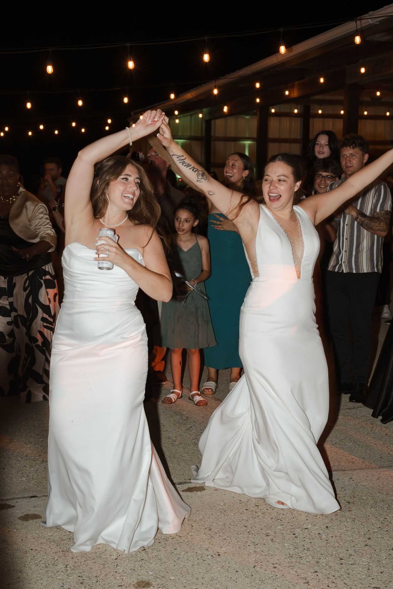 Two brides in white gowns dance joyfully at their wedding reception under string lights.