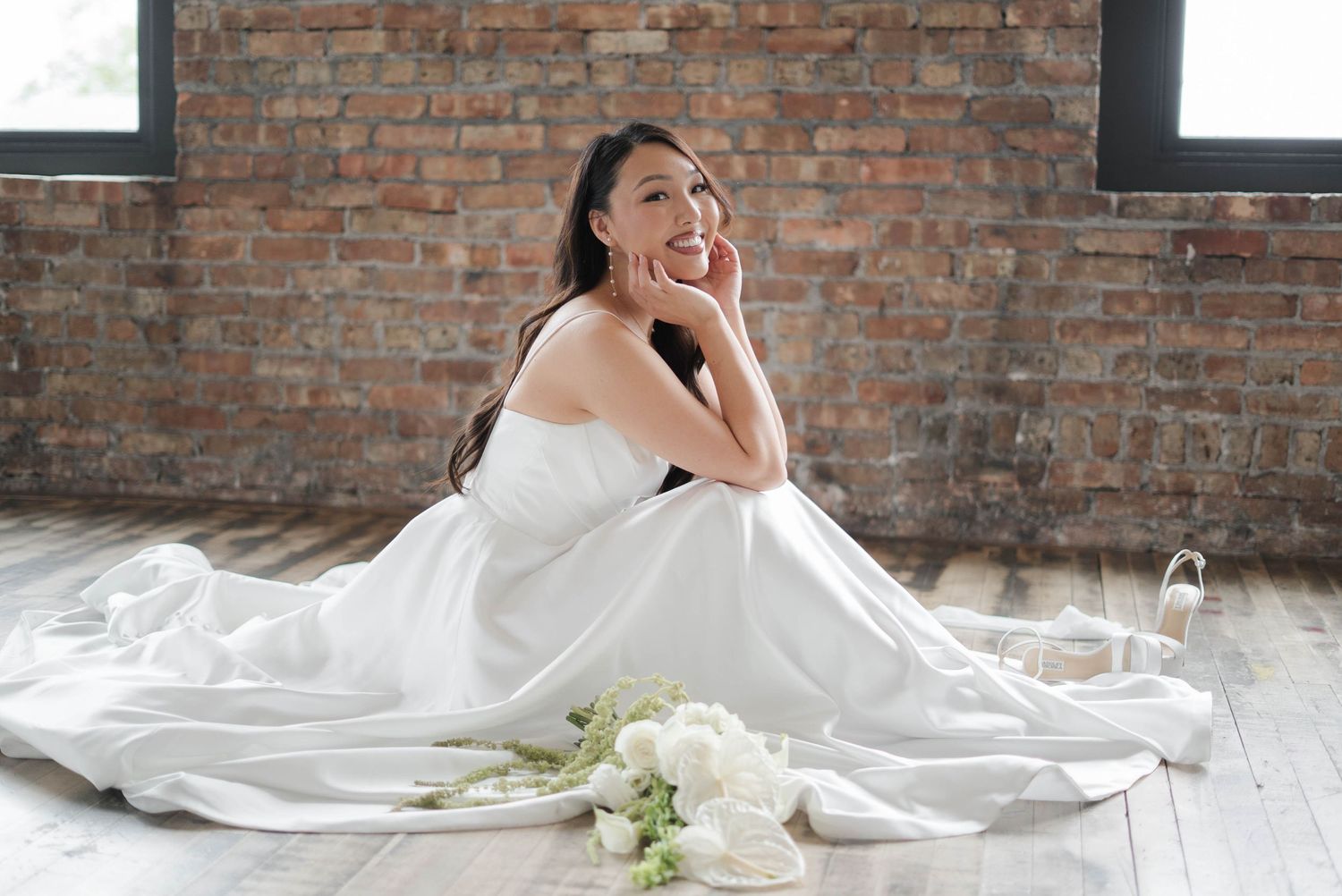 A bride in a white strapless gown sits gracefully on a wooden floor beside white flowers and heels.