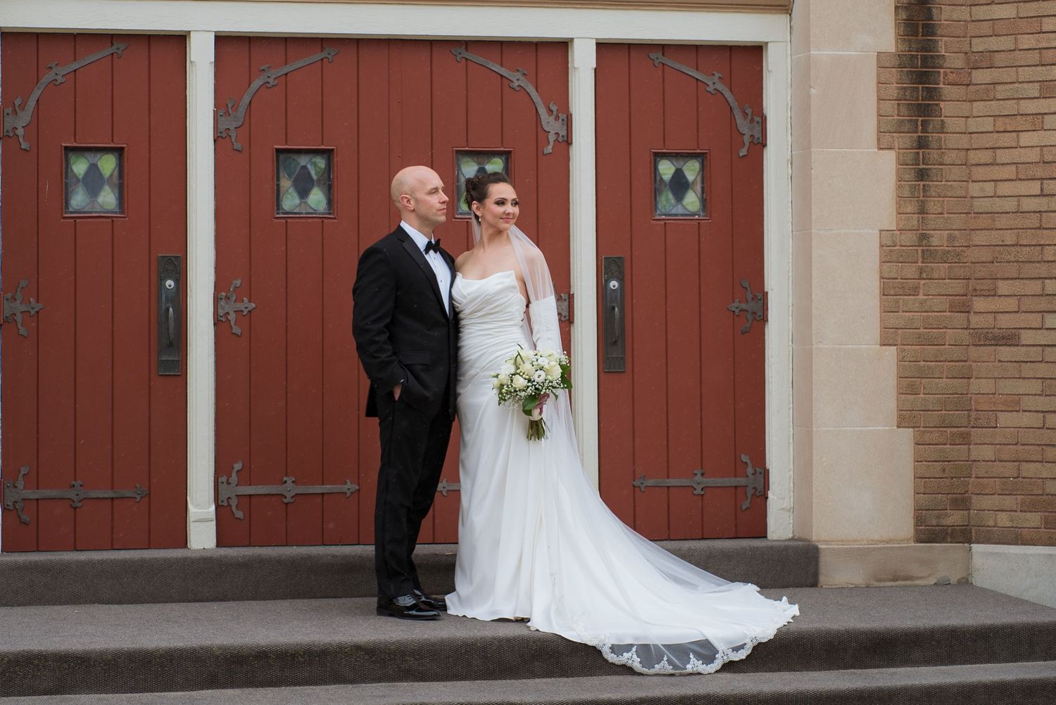 Wedding couple stands in front of brick building with red wooden doors during outdoor photo session.