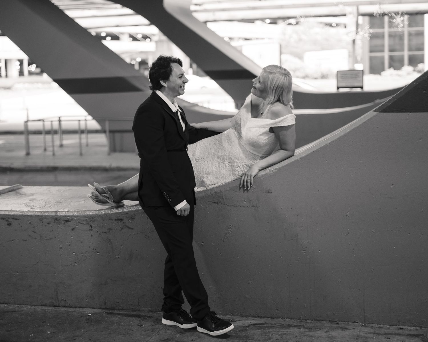 Black and white photo of a couple in wedding attire sharing a romantic moment under a bridge structure.
