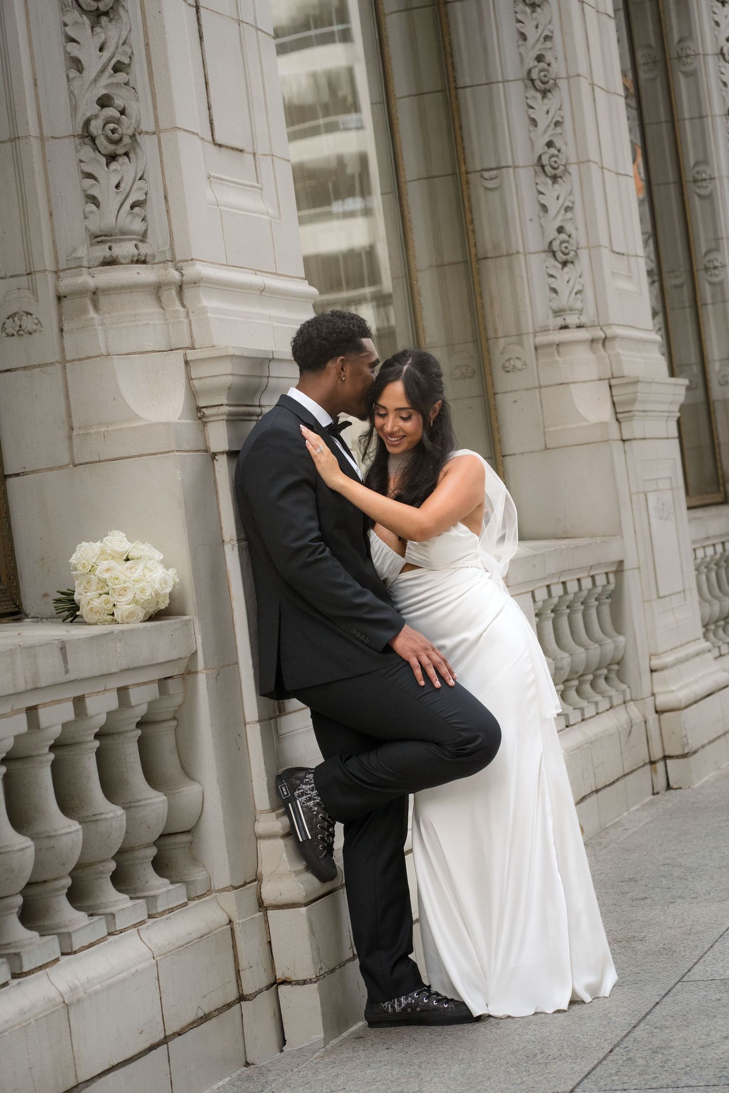 A groom in a black tuxedo embraces his bride in a white gown against an ornate stone building on their wedding day.