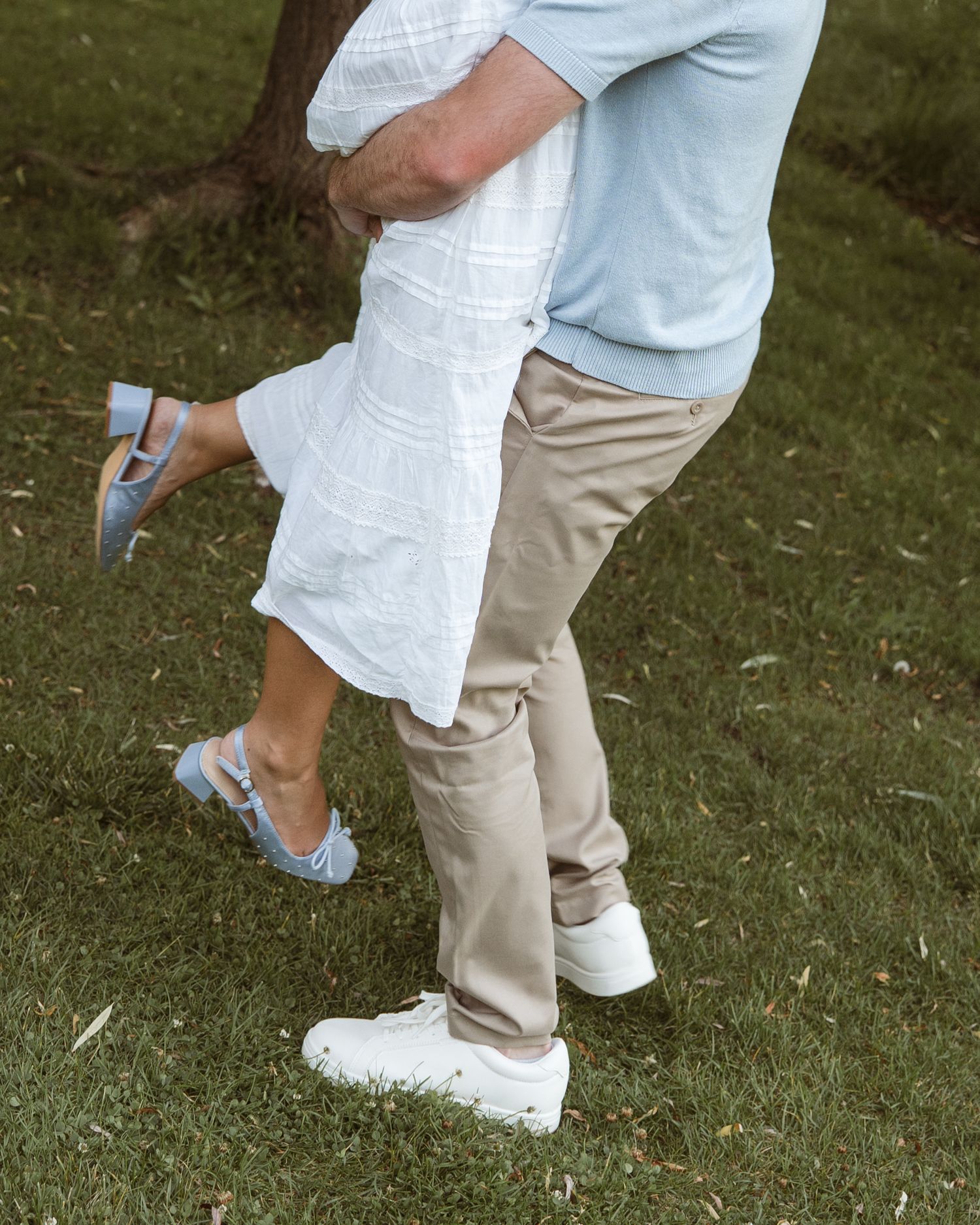 Couple embracing outdoors, woman in white dress and blue heels lifted by man in khakis and white sneakers on green grass.
