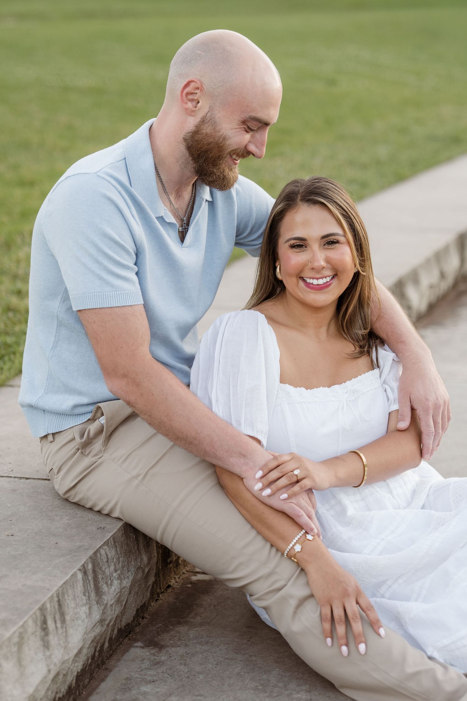 Couple sitting on stone steps outdoors, man in blue shirt embracing smiling woman in white dress.