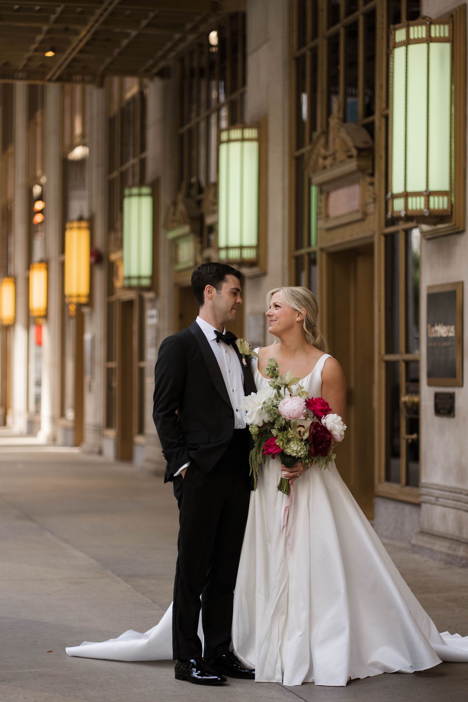 Bride and groom in wedding attire pose together in an elegant urban arcade with glowing lanterns.
