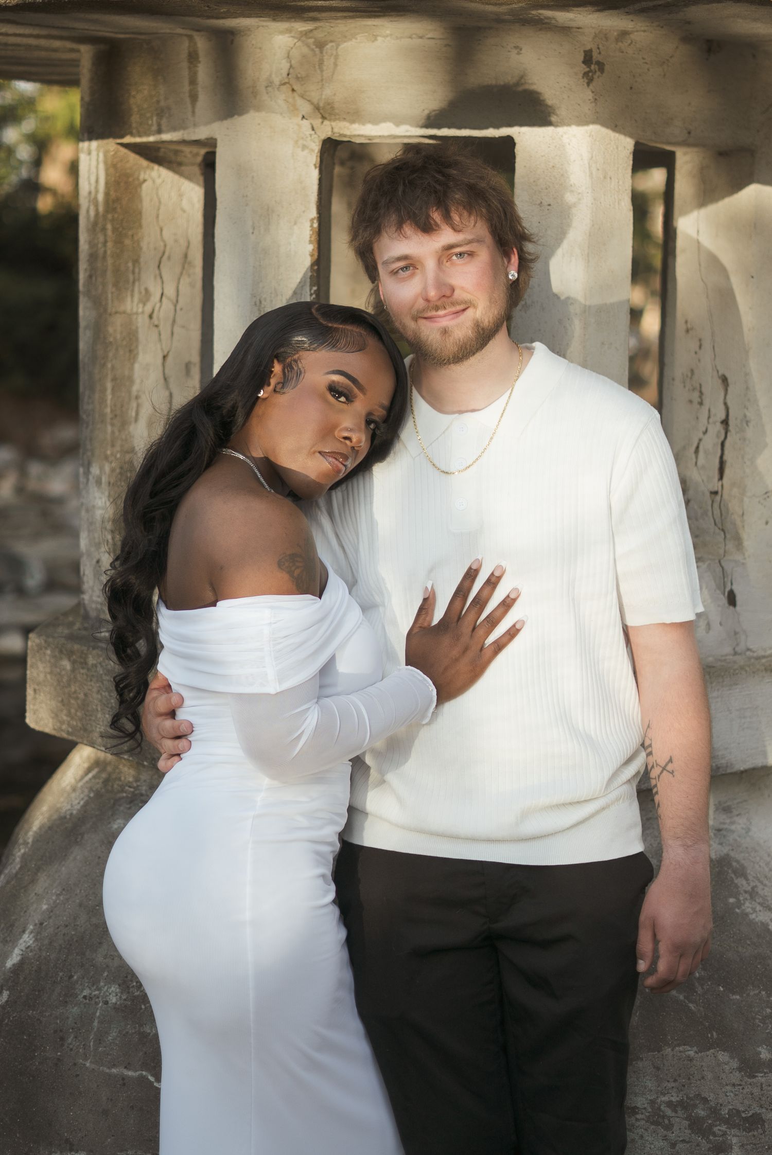 A couple in white outfits poses together outdoors, the woman in a fitted dress leaning against the man.