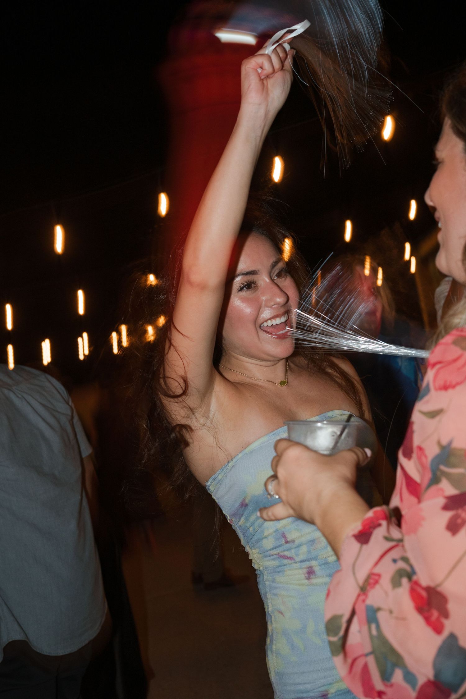 Woman dancing joyfully at a party, holding a drink and smiling, with festive lights glowing in the background.