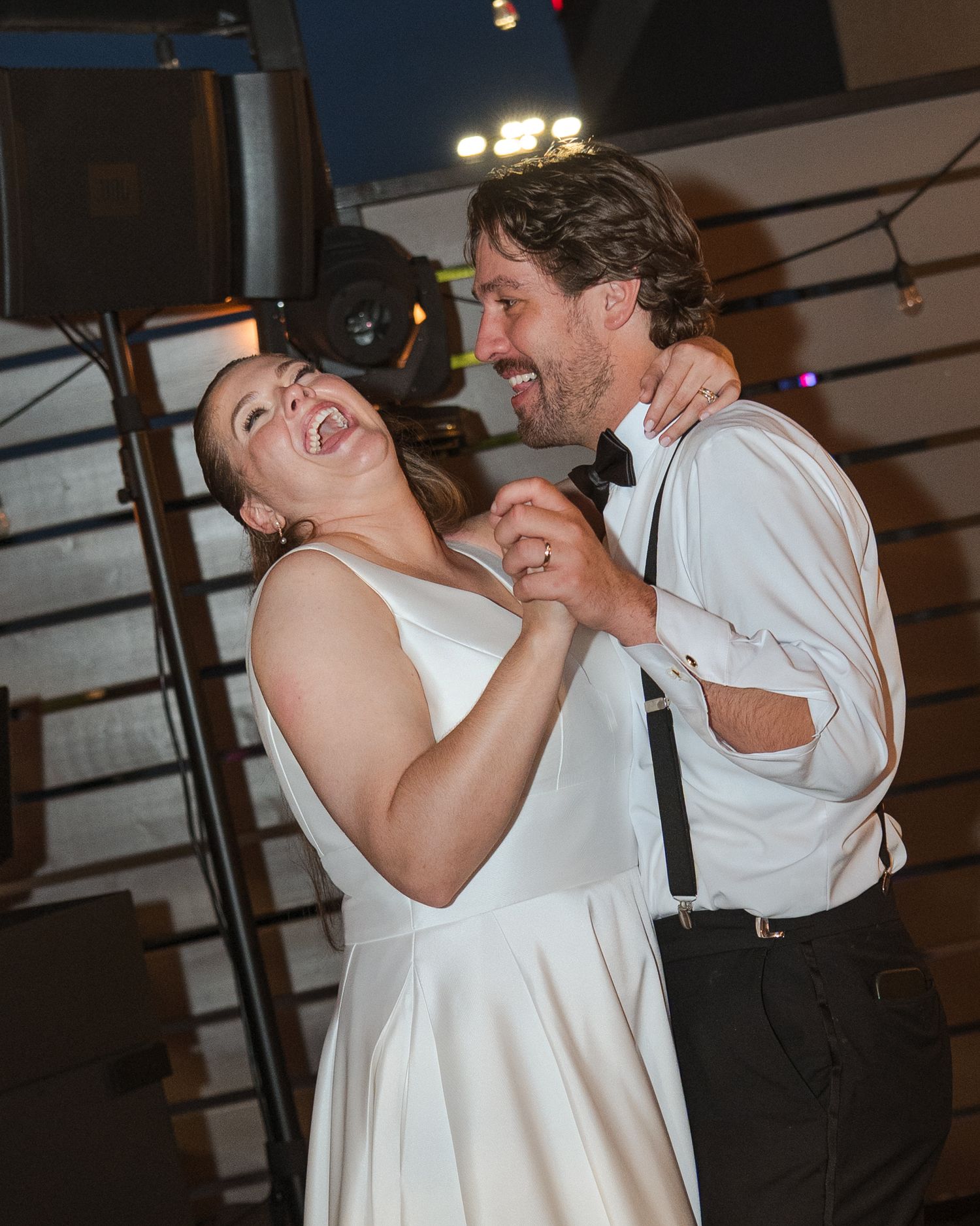 Joyful couple sharing first dance at wedding, bride in white gown laughing, groom in suspenders and bow tie smiling.