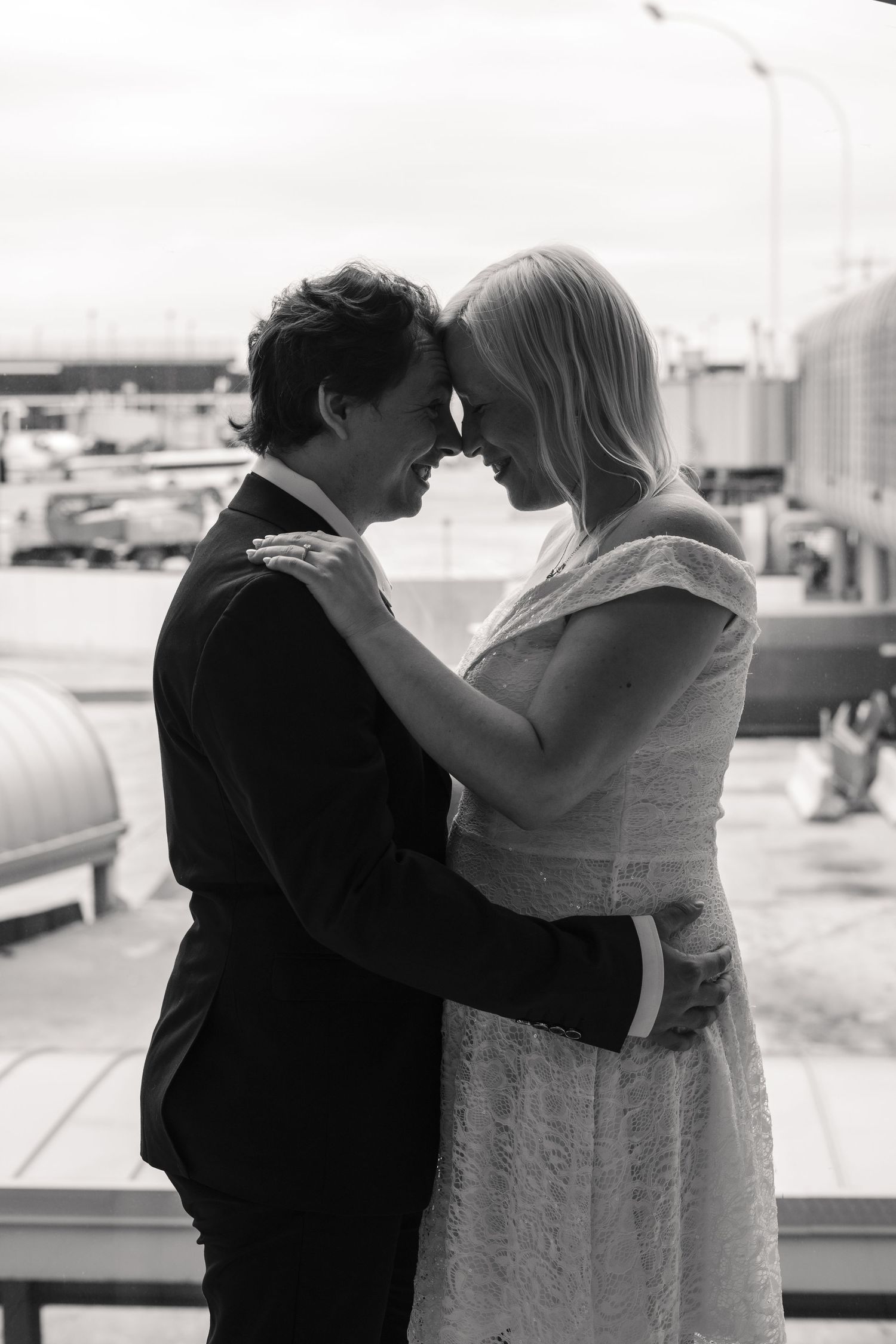 Black and white photo of a couple in wedding attire touching foreheads tenderly at an airport terminal.