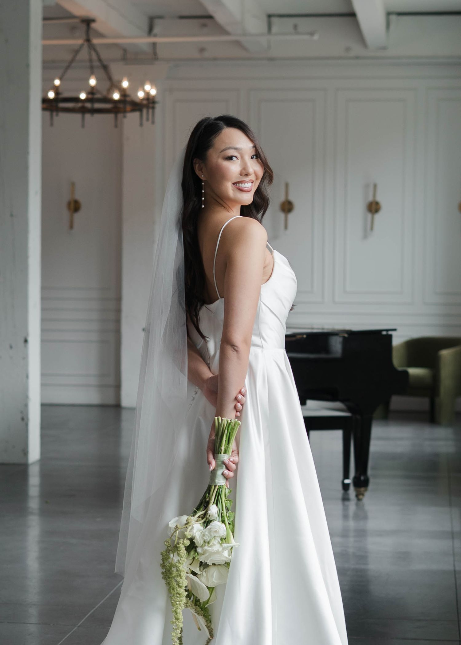 Bride in white satin gown with veil holds white bouquet, smiling in elegant venue with grand piano.