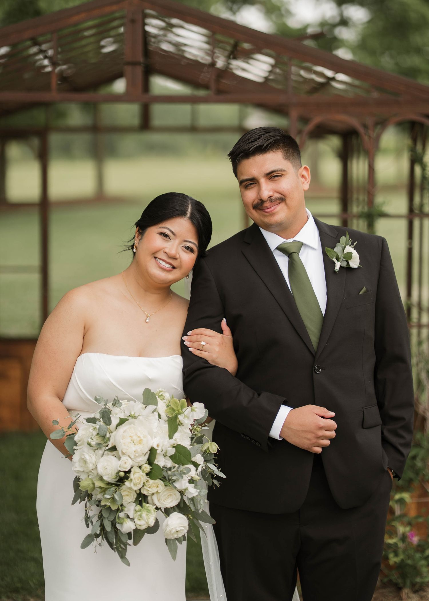 Bride and groom posing together outdoors, bride holding white bouquet, groom wearing sage green tie.