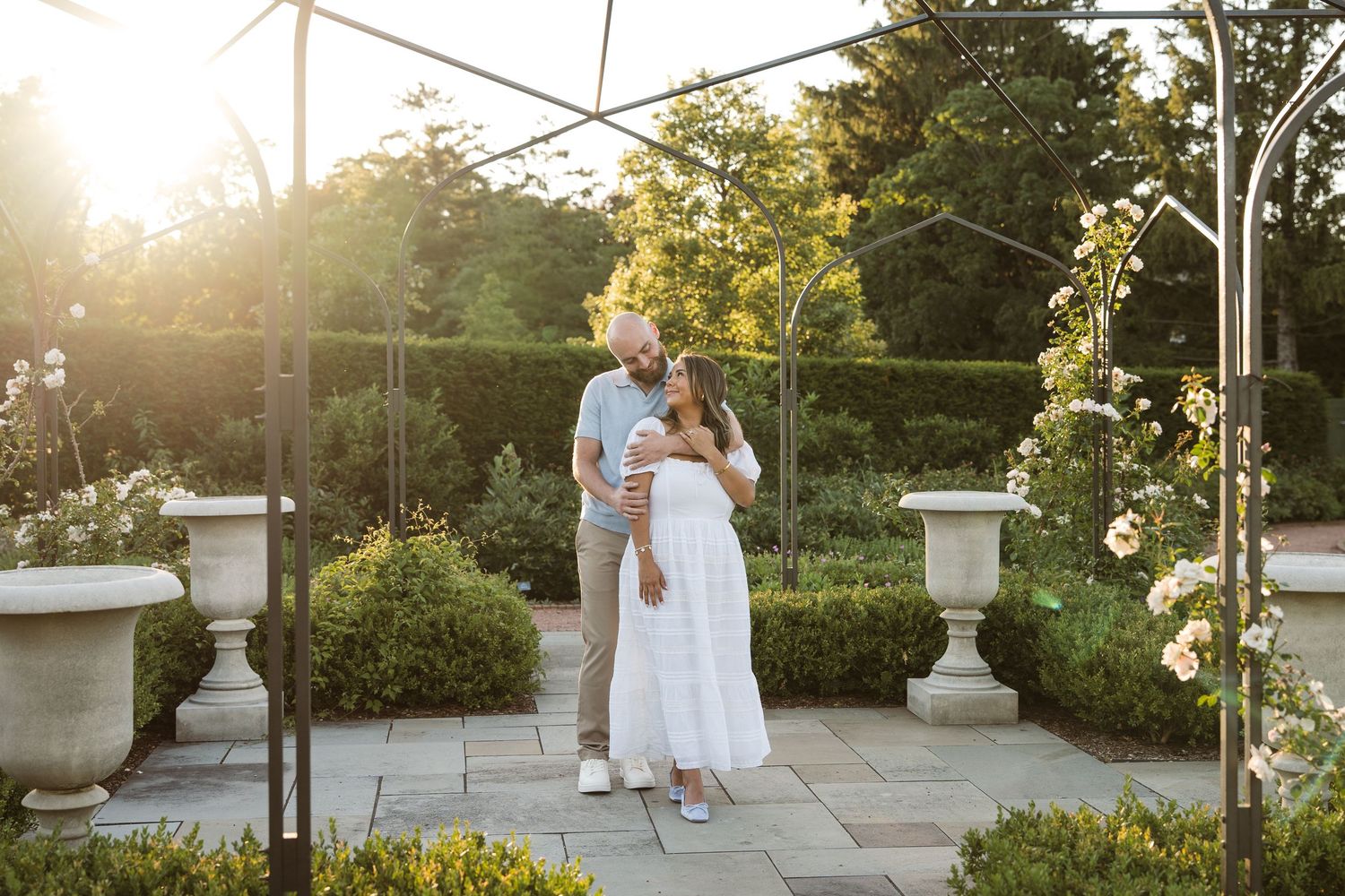 Couple embracing in a sunlit garden with lush hedges, stone urns, and a floral arch during a maternity photo session.