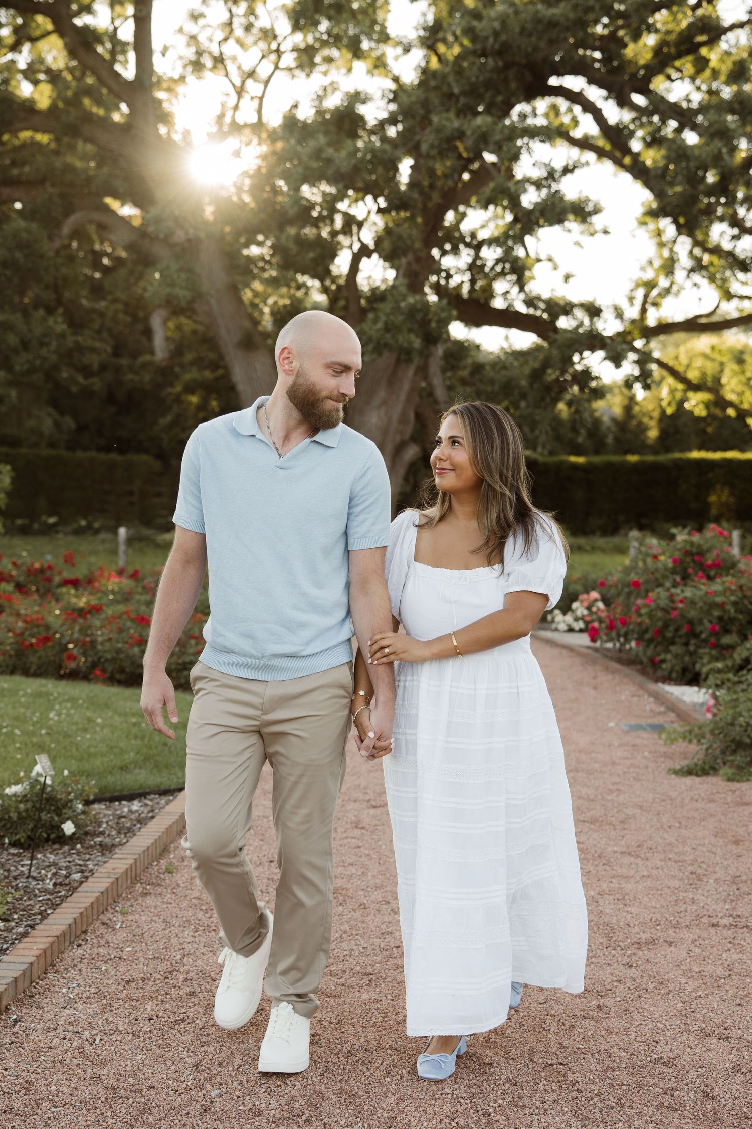Couple walking hand in hand through a sunlit garden path, man in blue shirt and woman in white dress.