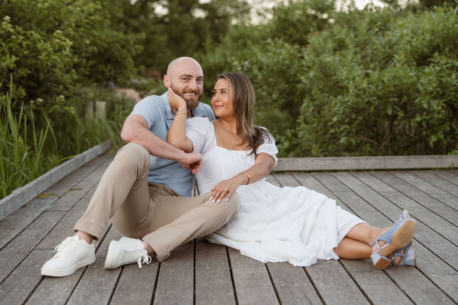 Couple sitting together on a wooden boardwalk surrounded by lush greenery, smiling and sharing a romantic moment.