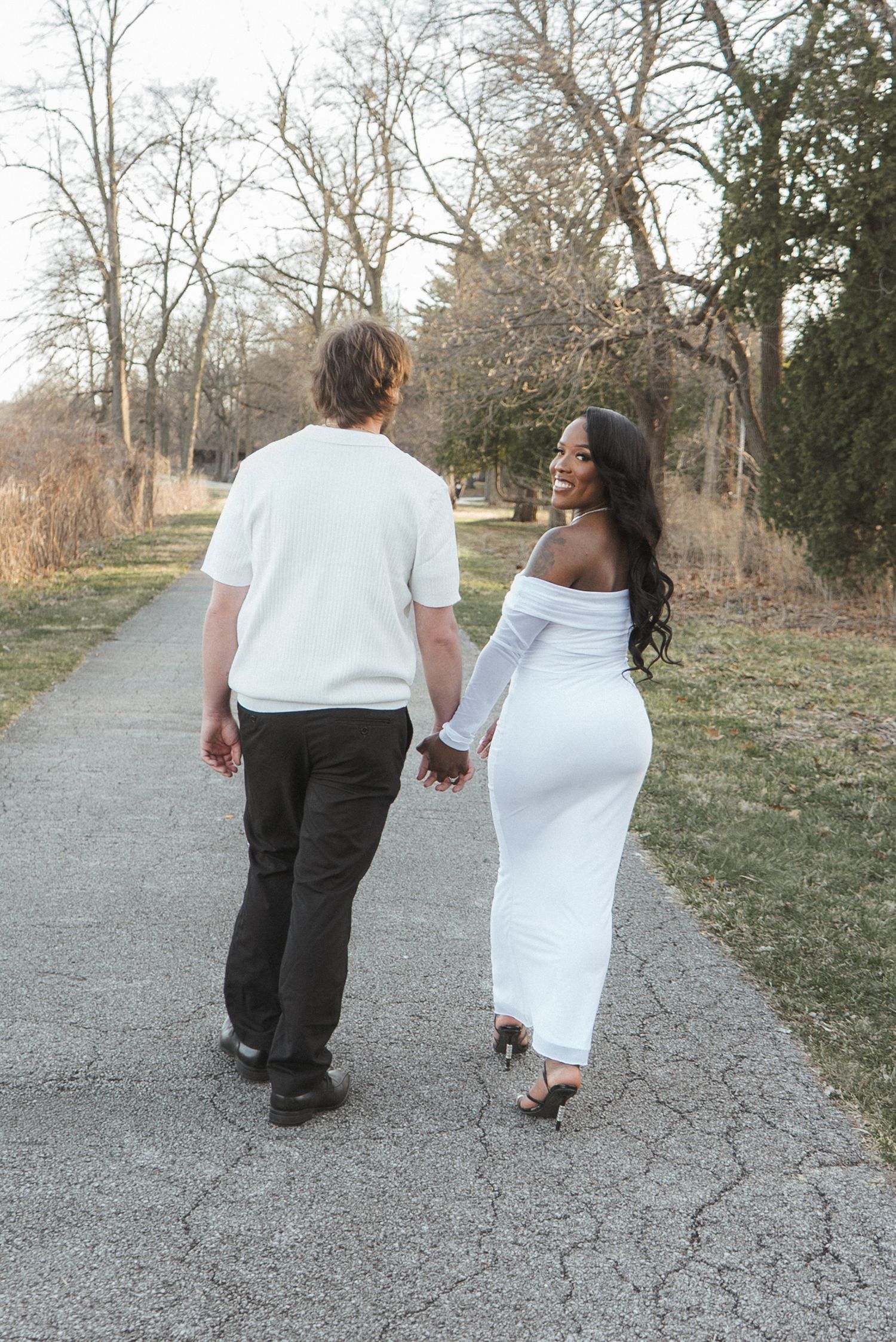 Couple holding hands walking on a path, woman in white off-shoulder dress smiling back, surrounded by bare trees.