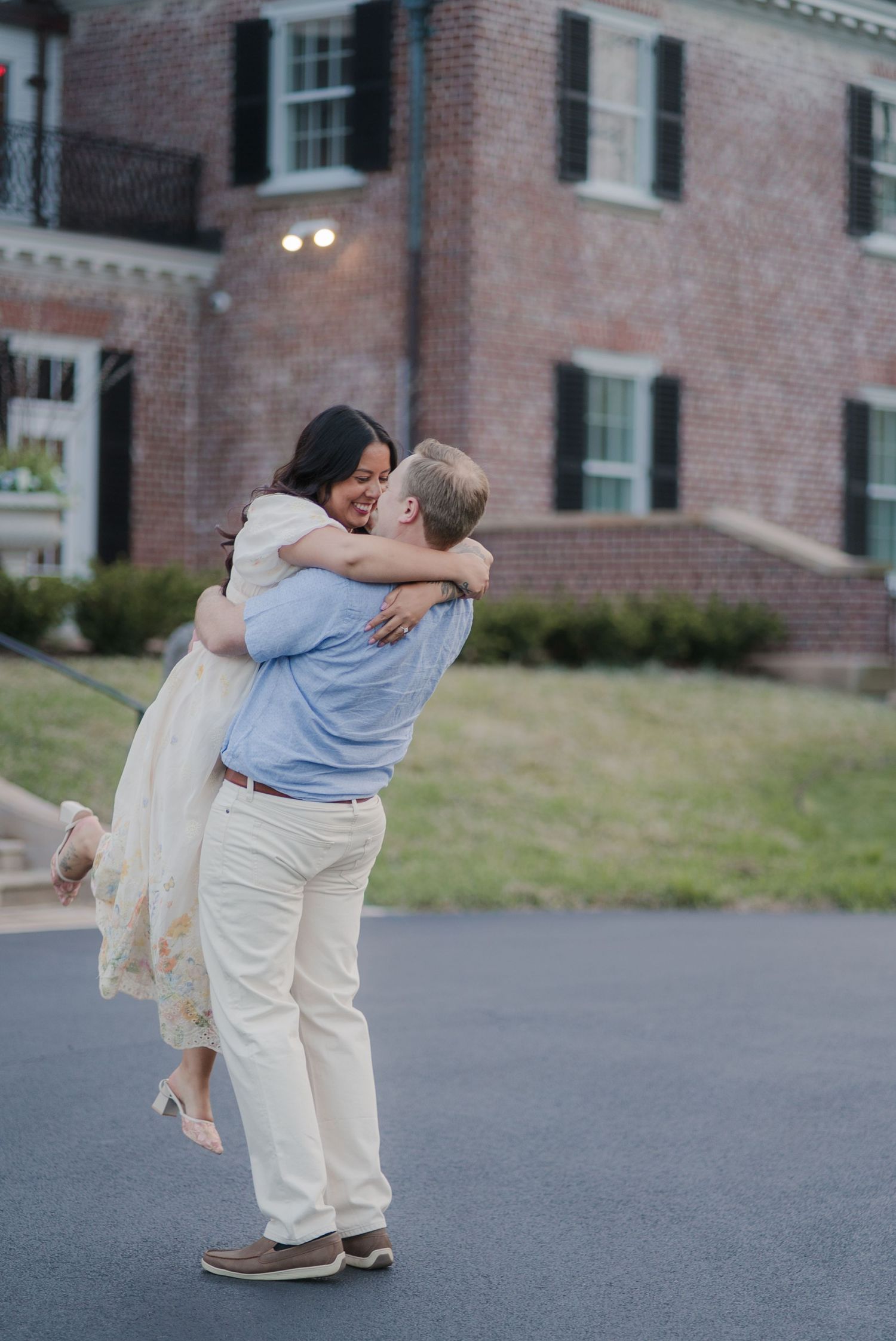 A man joyfully lifts a woman in a white dress outside a brick building during a romantic engagement photo session.