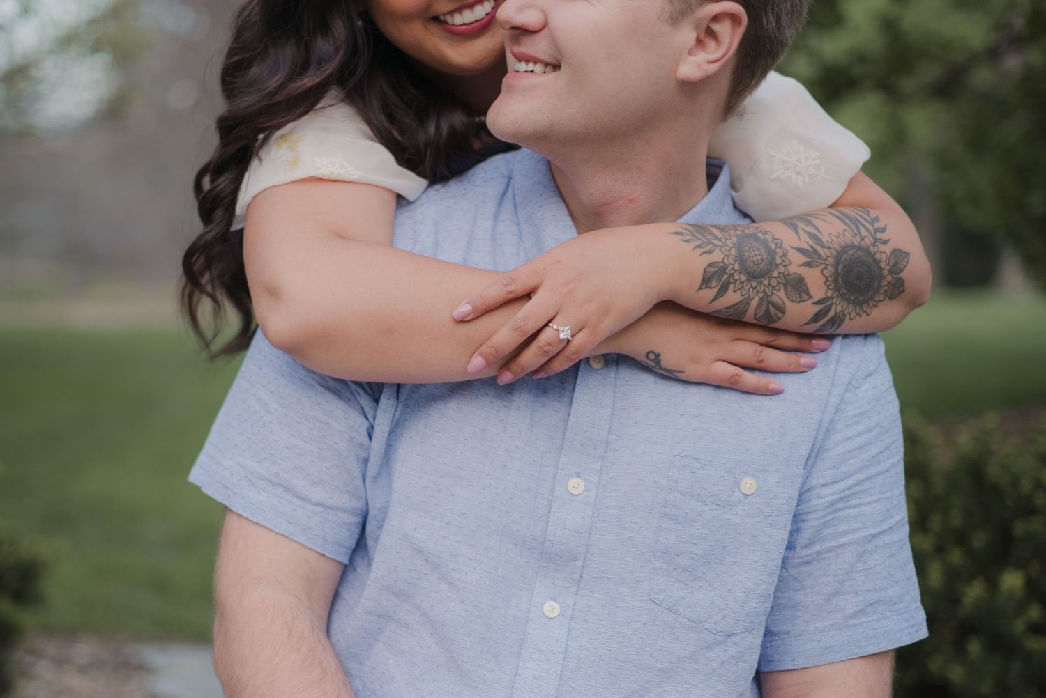 A smiling couple embraces outdoors, woman with dark curly hair hugging man in blue shirt with floral tattoo on arm.
