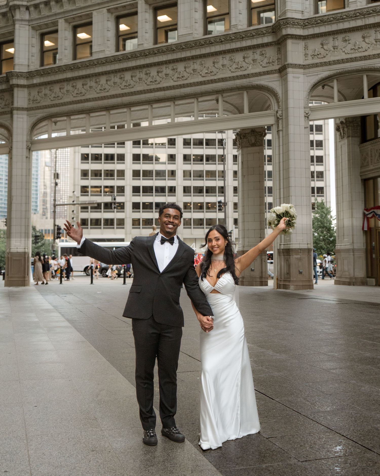Joyful interracial newlywed couple celebrating in front of grand Chicago architecture, bride in white gown holding bouquet.