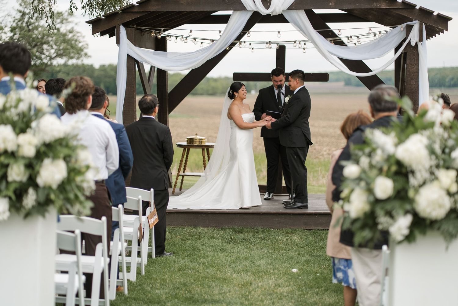 Outdoor wedding ceremony under a wooden gazebo with white draping, guests seated in white chairs on a green lawn.