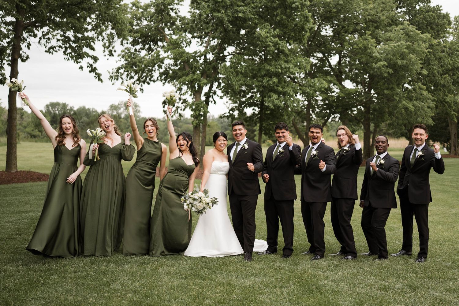 Wedding party in olive green and black attire celebrating outdoors on lush green lawn with trees in background.