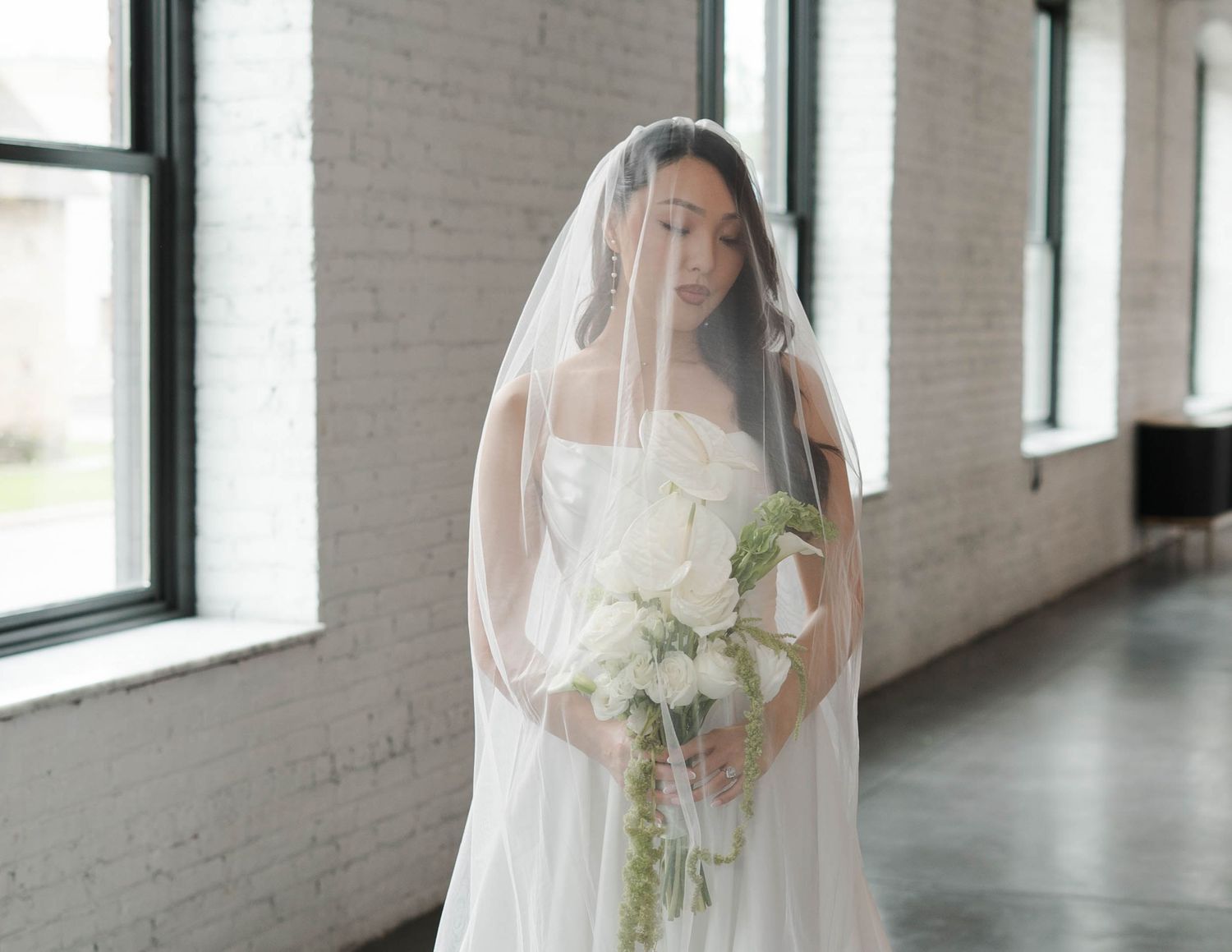 Bride in white gown and veil holding white floral bouquet standing in bright industrial venue with brick walls.