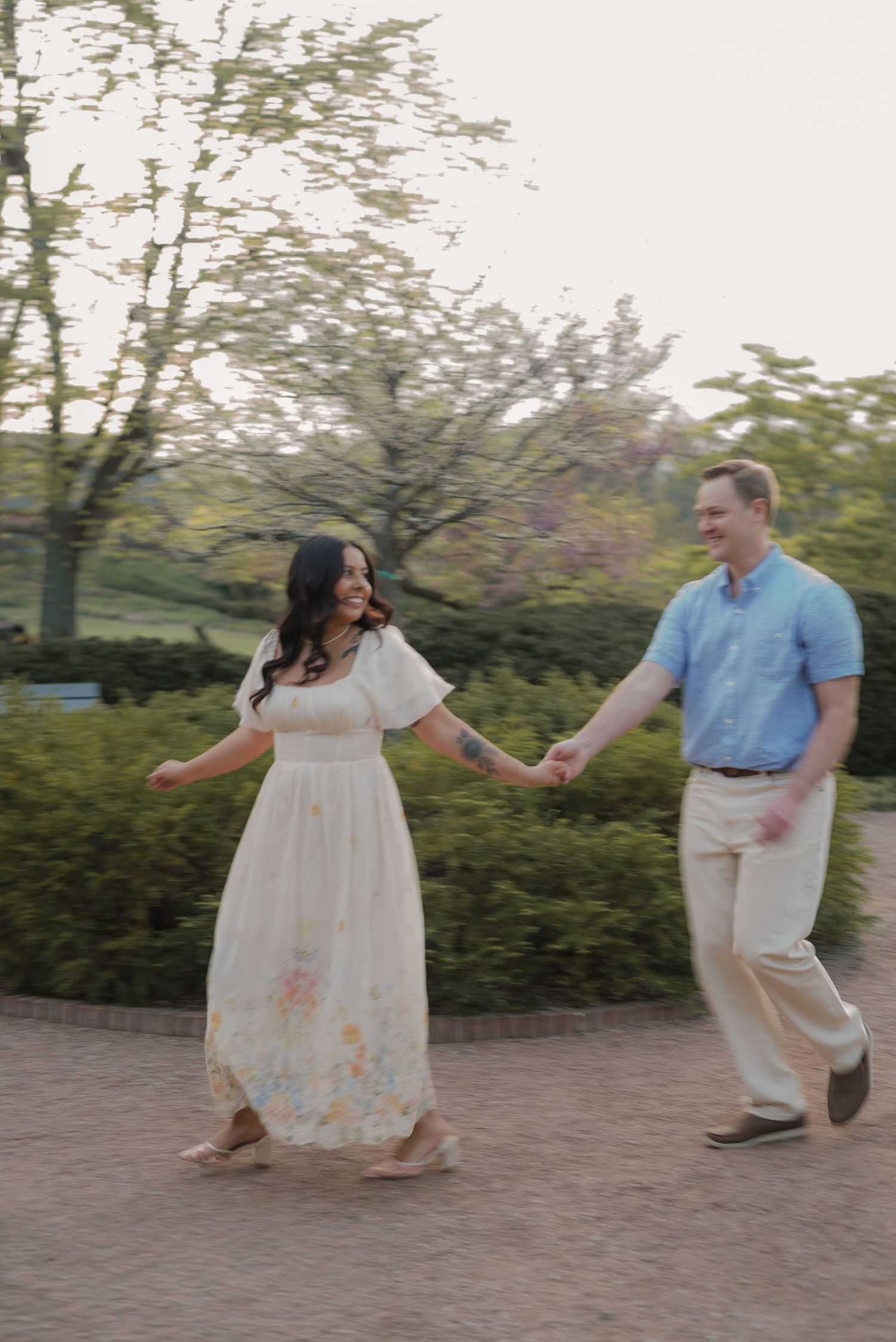 A couple holds hands and walks together in a garden, smiling, as the woman twirls in a floral maxi dress.