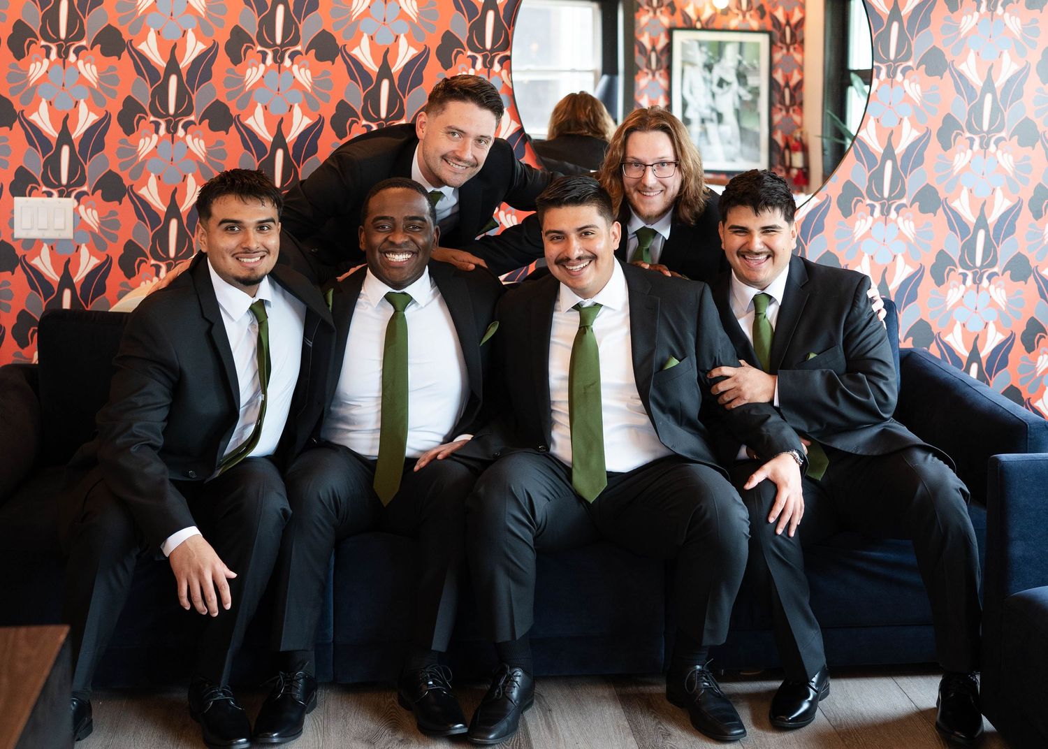 Six groomsmen in dark suits with green ties smiling together on a blue couch in front of a decorative wall.