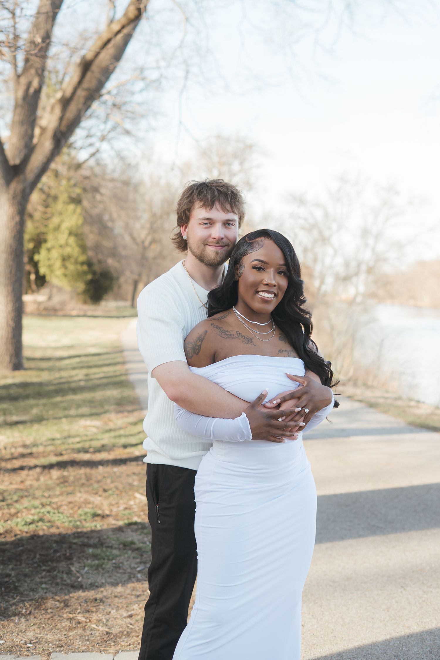 Couple embracing outdoors in white outfits near a lake, smiling warmly in a romantic engagement photo.