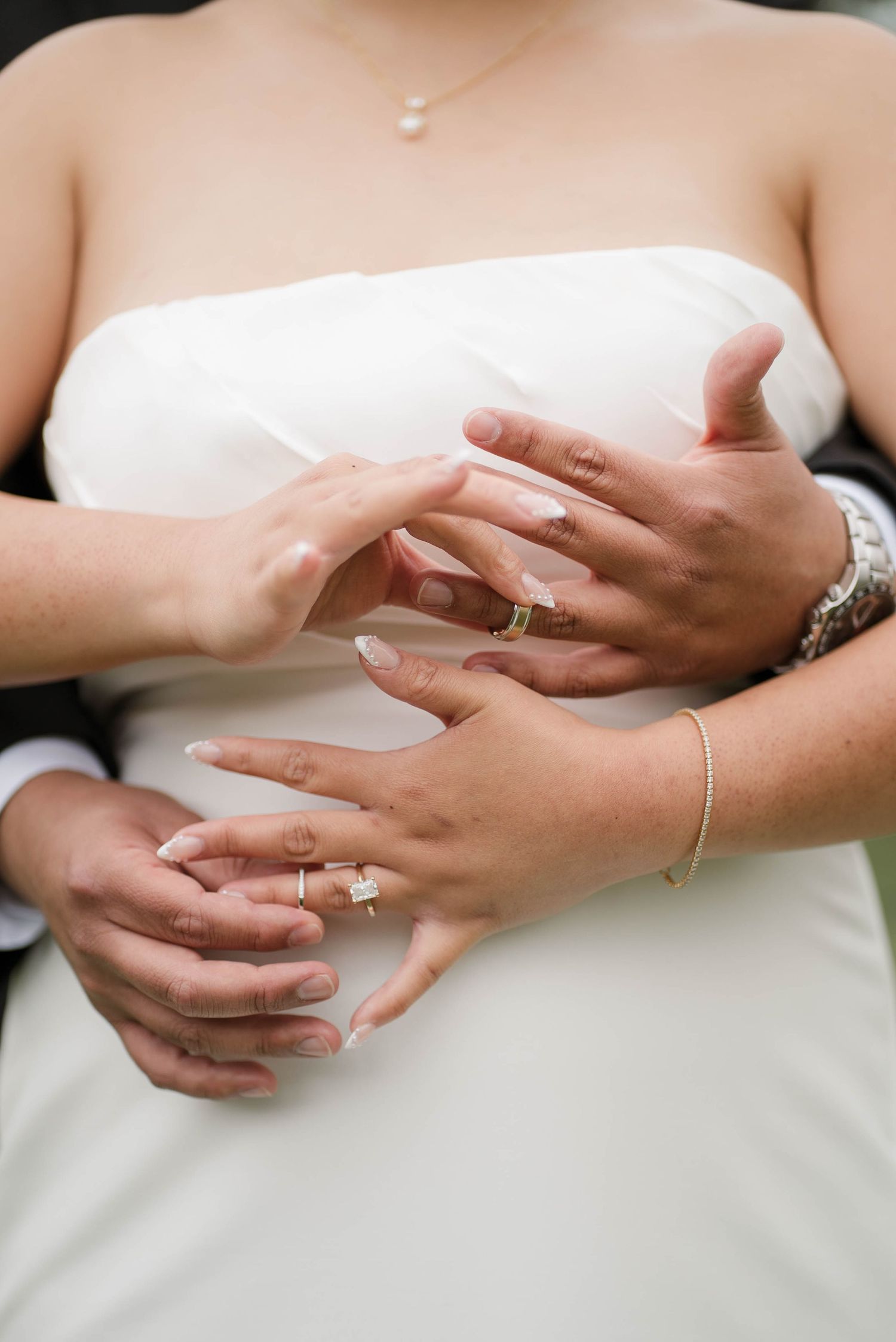 Close-up of bride and groom hands showing wedding rings, bracelet, and engagement ring on white wedding dress.