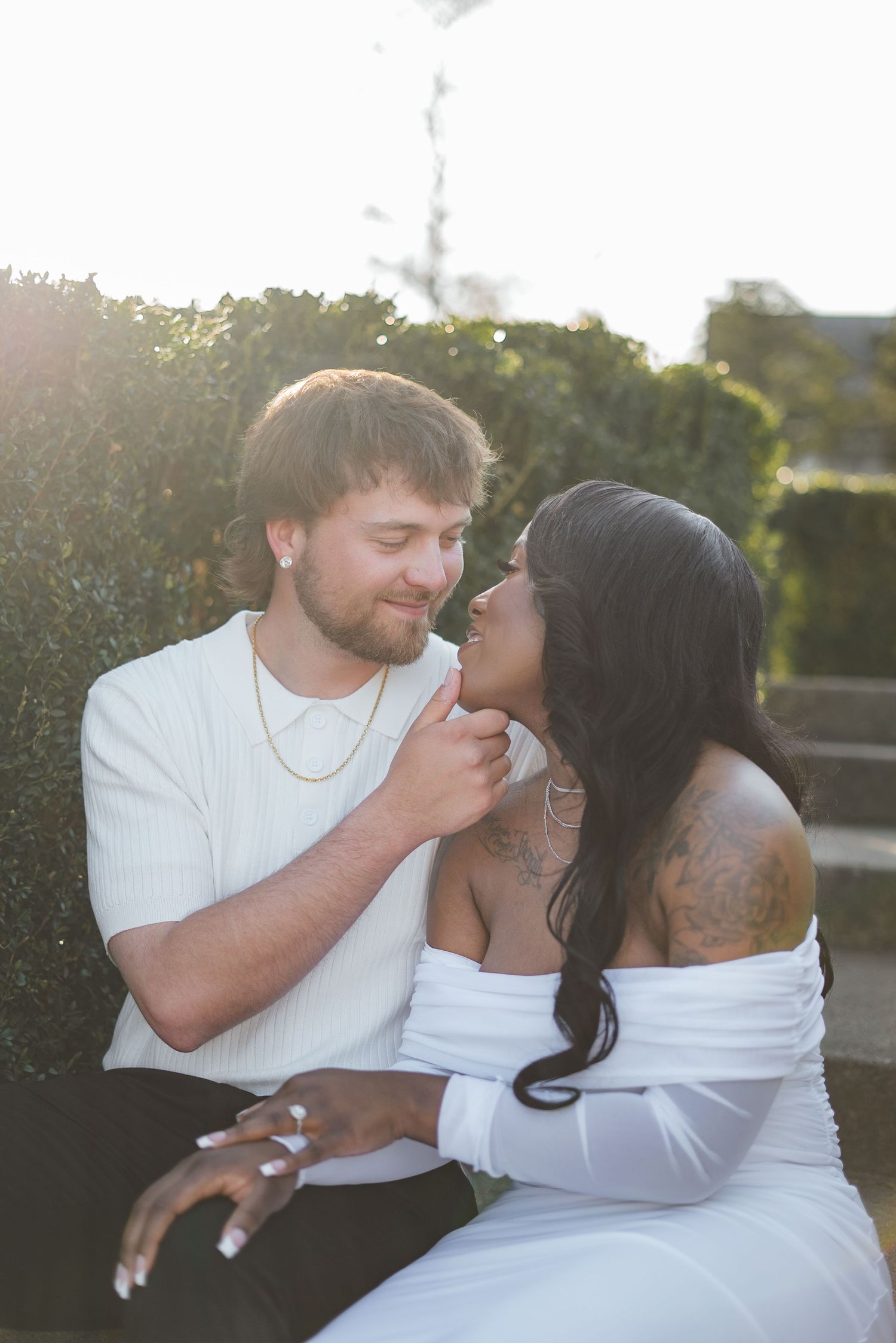 A couple shares a tender moment outdoors, man in white shirt touching womans chin, woman in off-shoulder dress with tattoos.