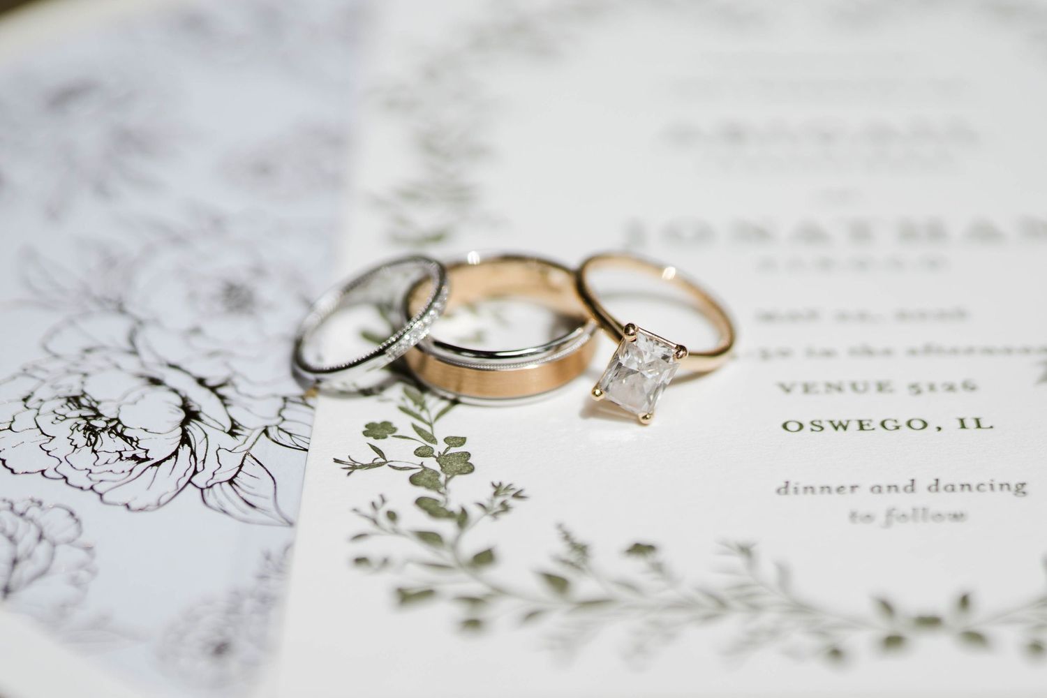 Wedding rings and diamond engagement ring displayed on a floral invitation in Oswego, IL.