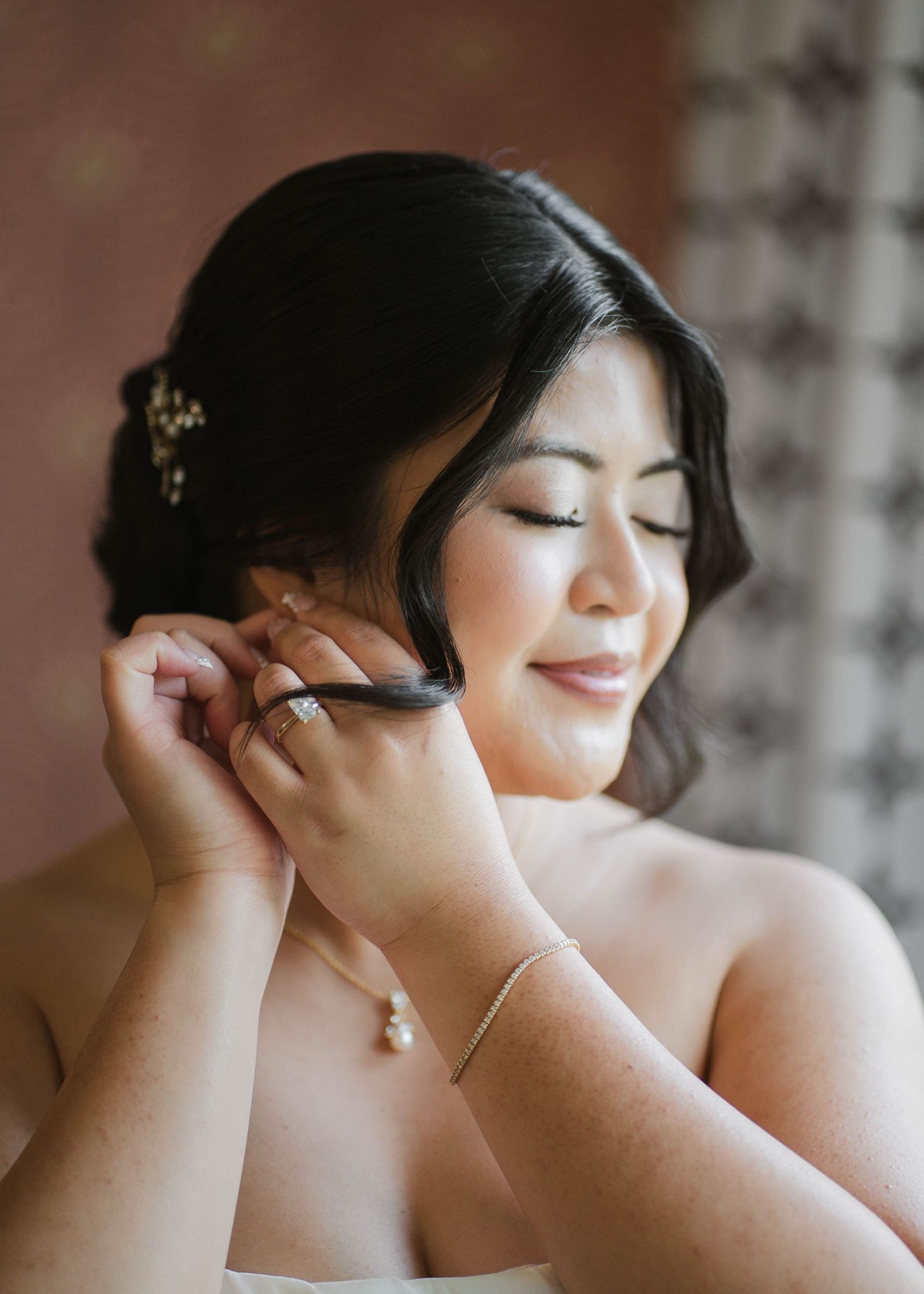 Bride putting on earrings with eyes closed, wearing pearl necklace and bracelet, with floral hair accessories.