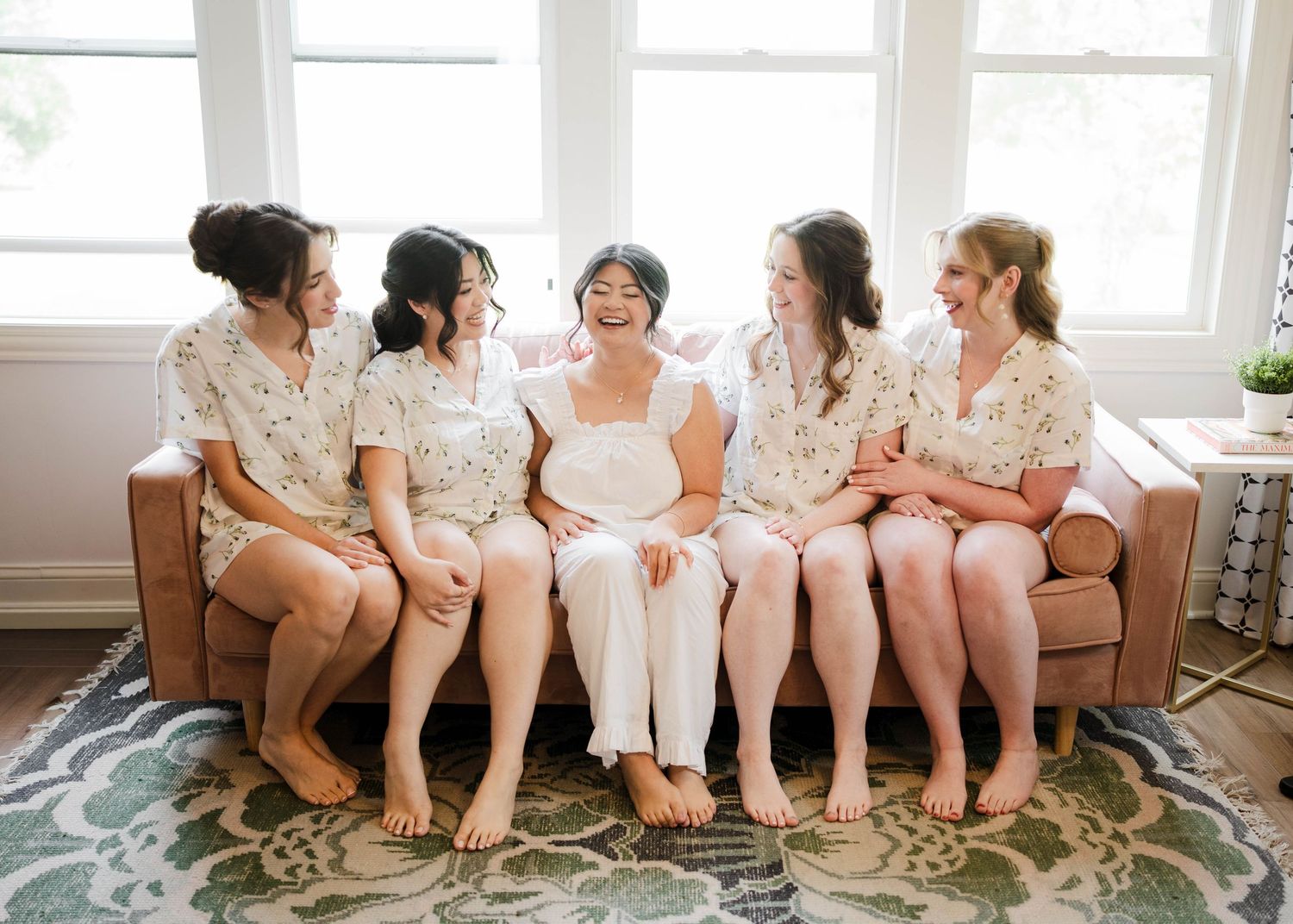 Bride and bridesmaids laughing together on a couch in matching floral robes during wedding morning preparations.