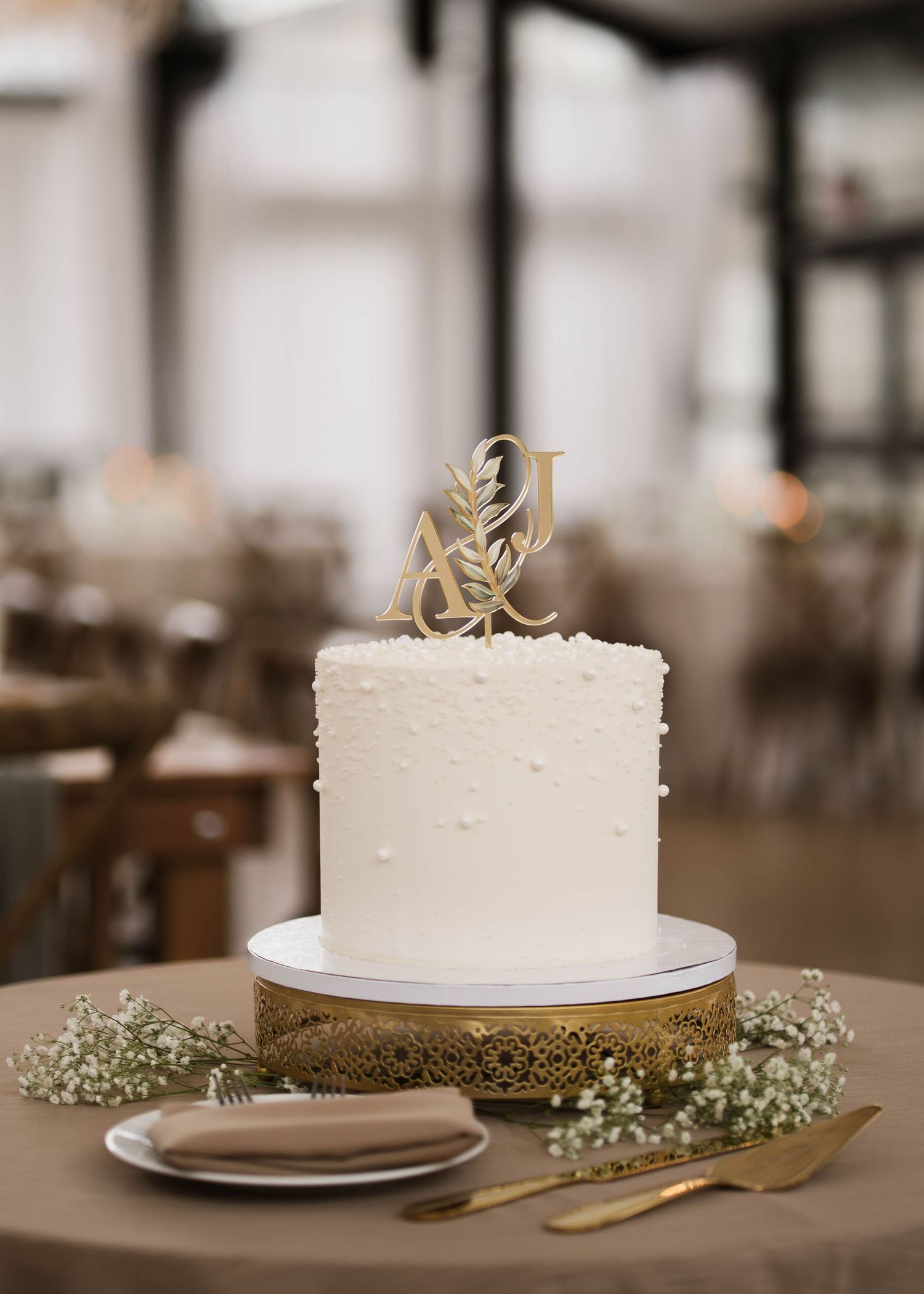 Single-tier white wedding cake with gold monogram topper on ornate gold cake stand.