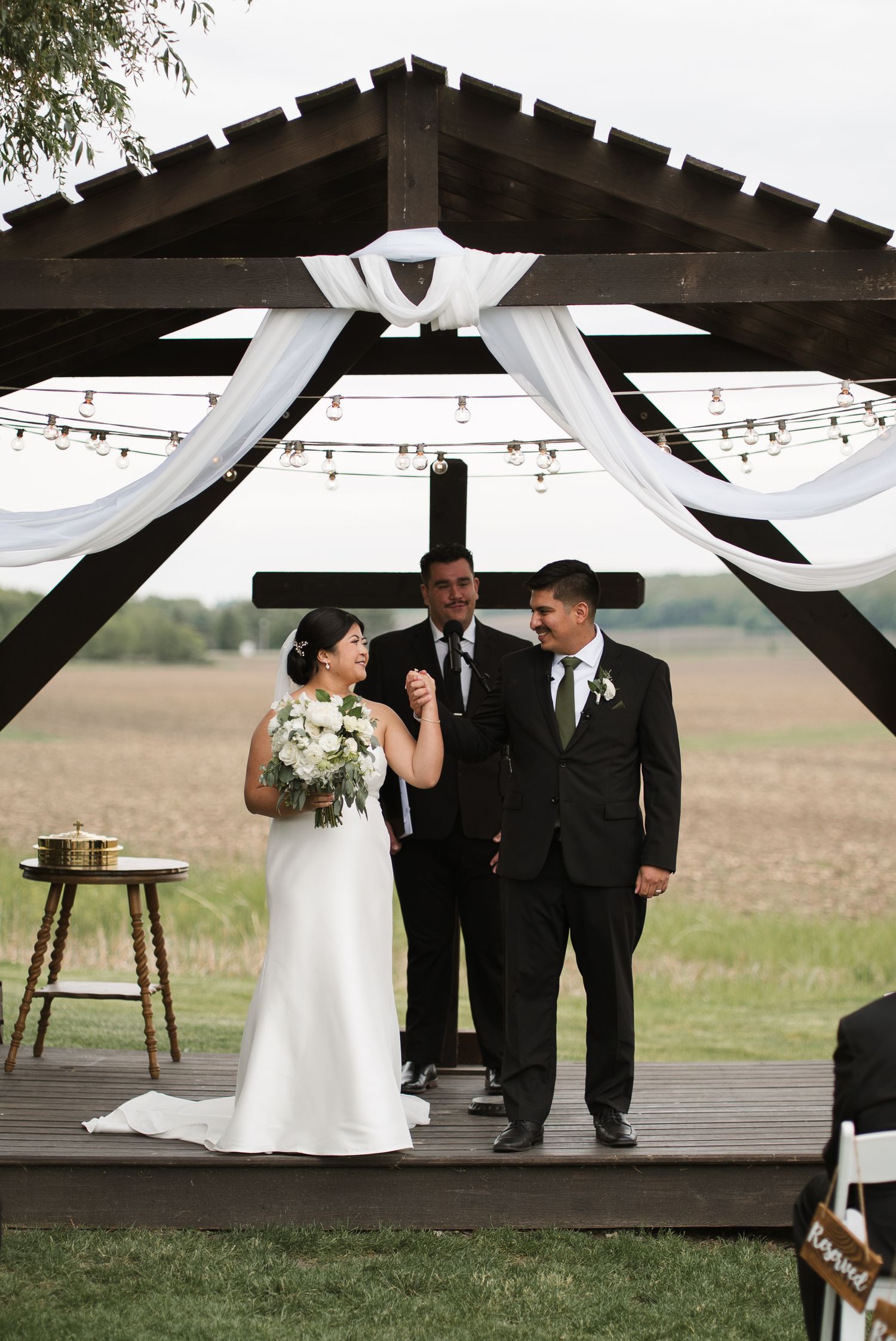 Bride and groom holding hands at outdoor wedding ceremony under rustic wooden pergola with white draping and string lights.