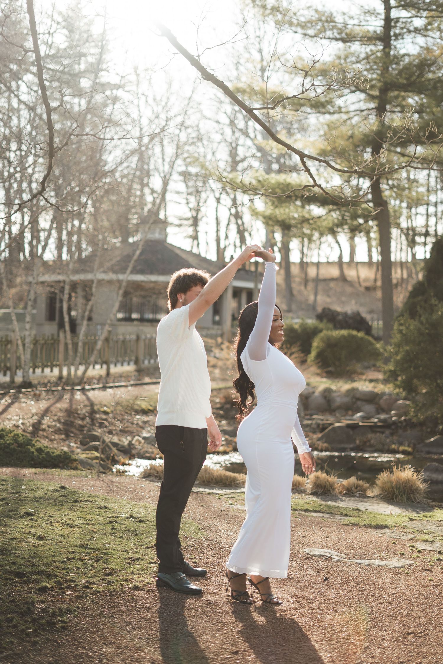 Couple dancing outdoors in white outfits, man spinning pregnant woman on a sunny garden path surrounded by trees.