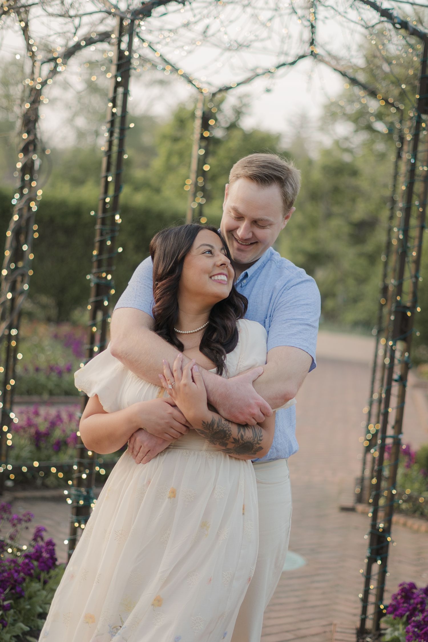 A couple embraces and laughs under a fairy light arch surrounded by purple flowers in a romantic garden setting.