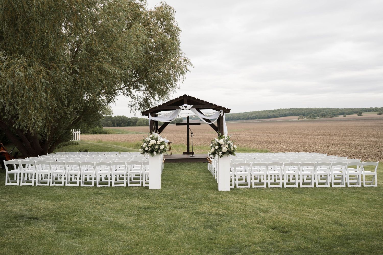 Elegant outdoor wedding ceremony setup with white chairs, floral arrangements, and a decorated pergola altar in a scenic field.