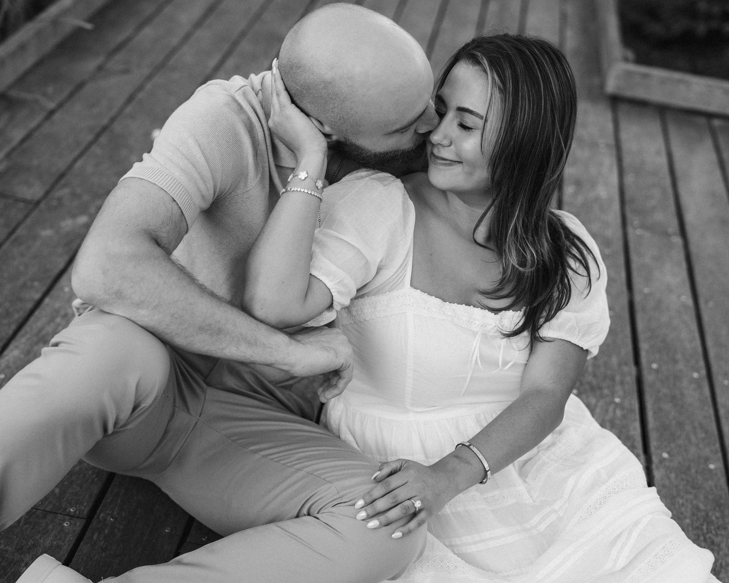 Black and white photo of a couple sitting on wooden deck, man kissing womans cheek as she smiles warmly.