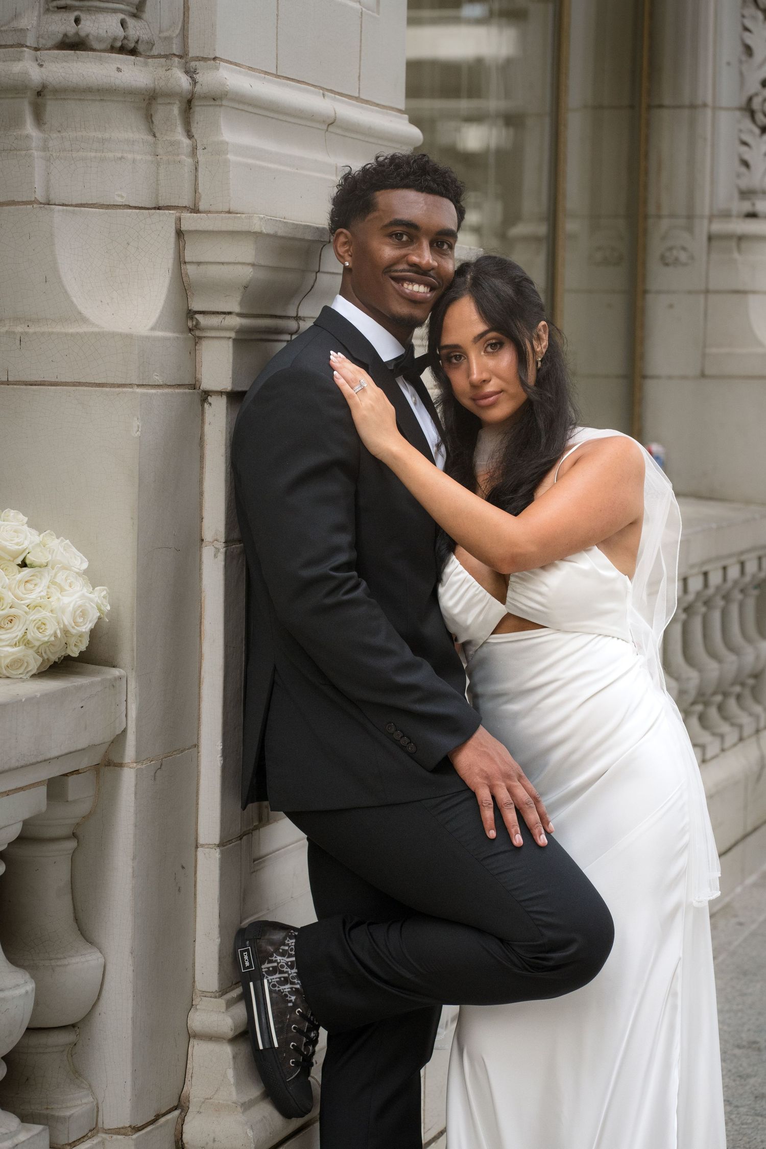 Interracial wedding couple posing elegantly against ornate stone building, bride in white gown.