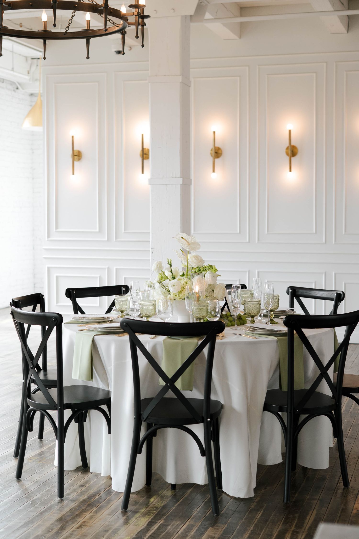 Elegant round dining table with white linens, black cross-back chairs, floral centerpiece, and gold wall sconces in a bright venue.