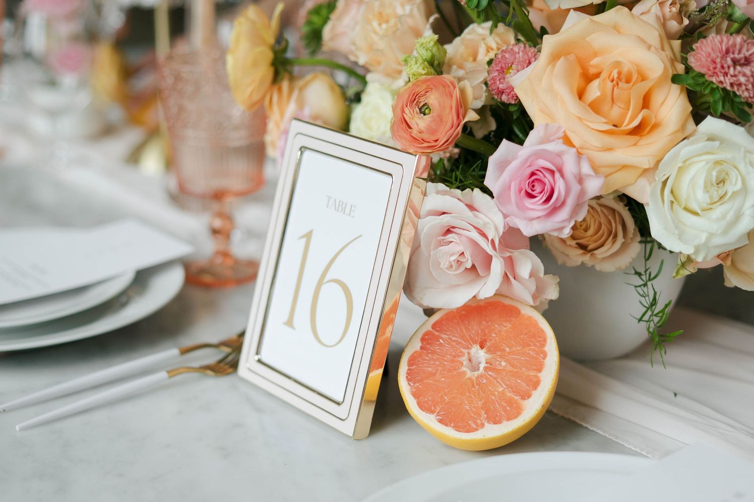 Elegant wedding table setting with a gold table number 16 card, pastel floral centerpiece, and a halved grapefruit.