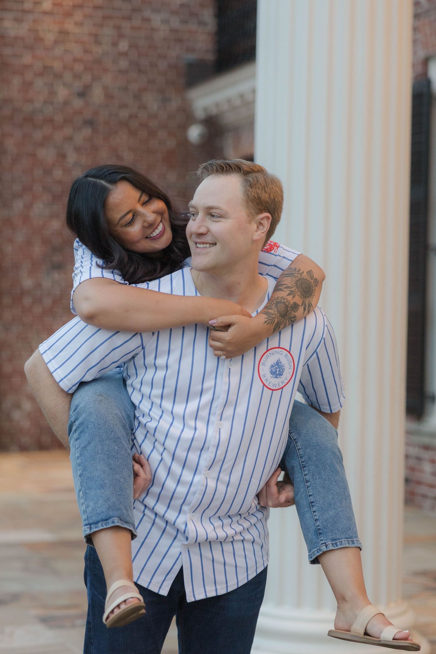Couple in matching baseball jerseys sharing a piggyback ride, laughing outdoors near brick building.