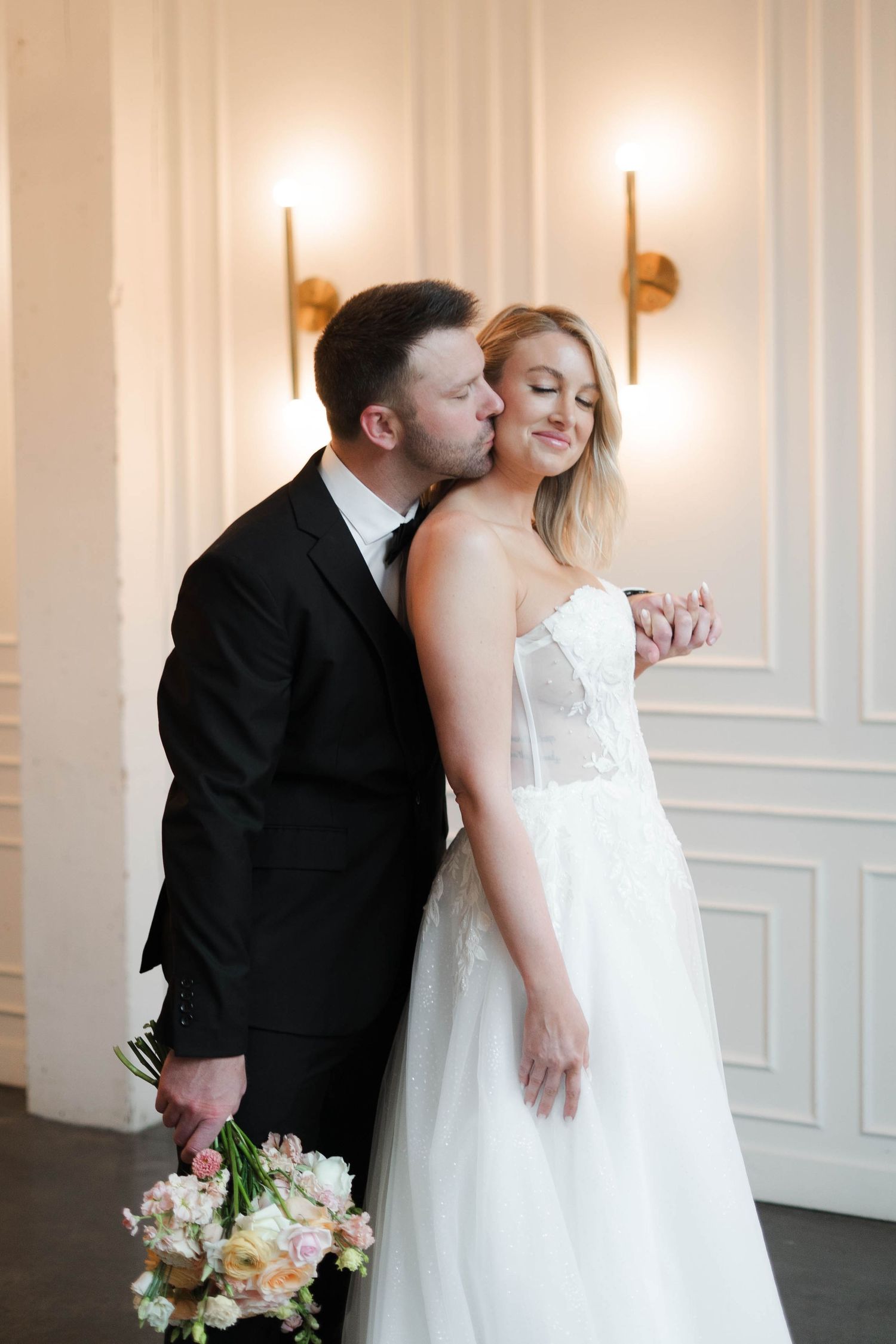 Groom in black tuxedo kissing bride in white strapless wedding gown holding floral bouquet indoors.