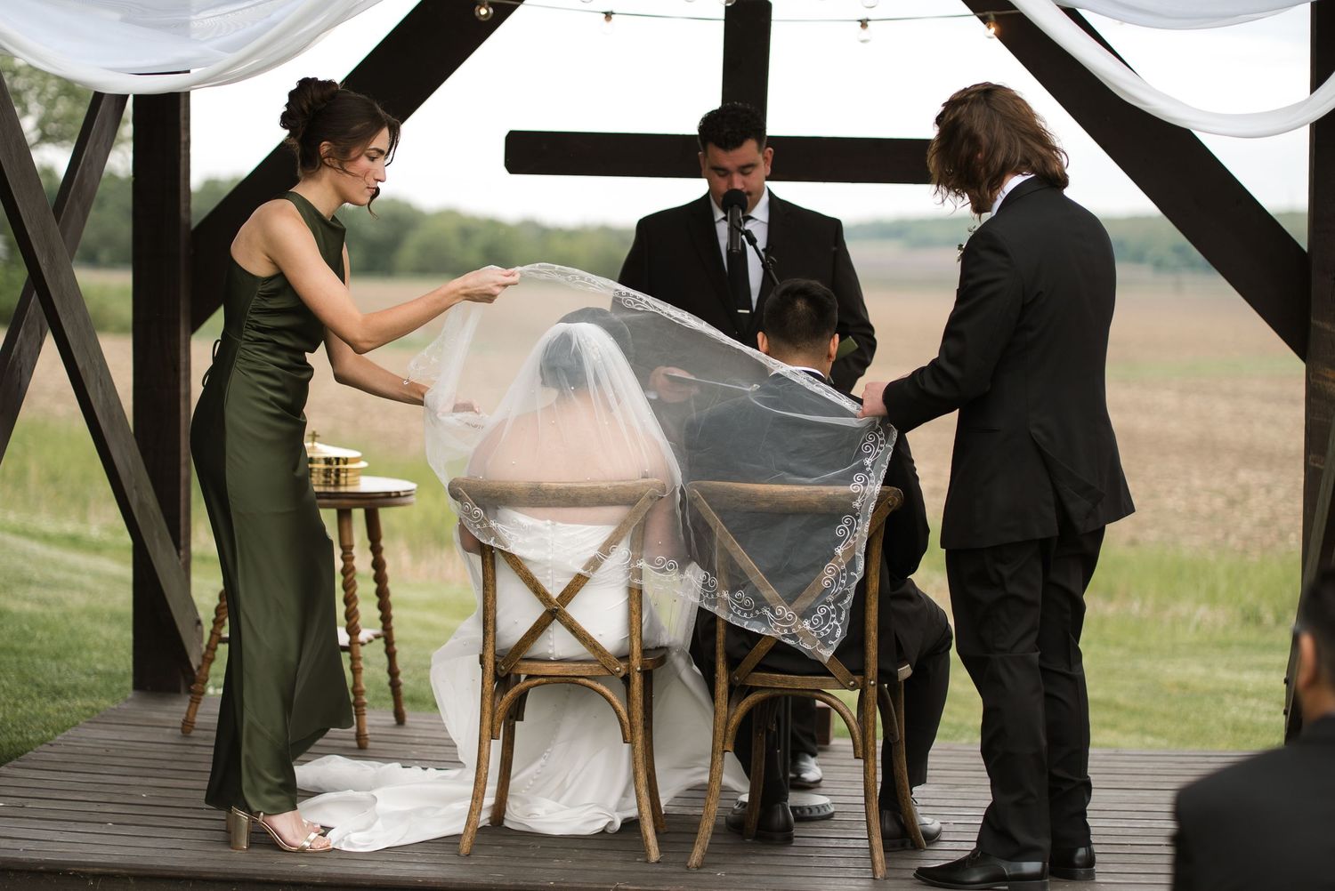 Outdoor wedding ceremony with bride seated as bridesmaid lifts veil, officiant and guests stand under wooden pergola.