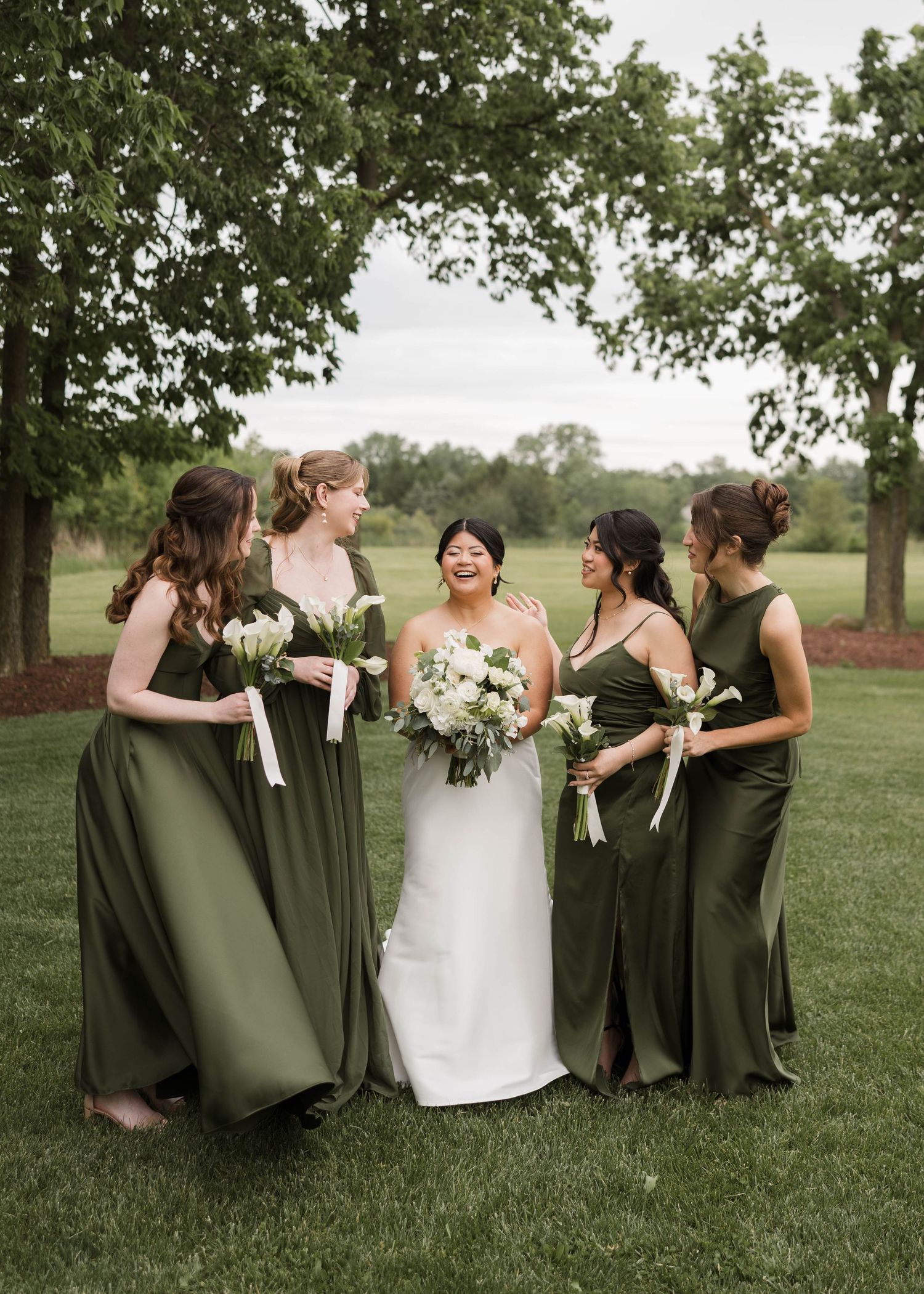 Bride and four bridesmaids in elegant olive green gowns laughing together on a lush green lawn.