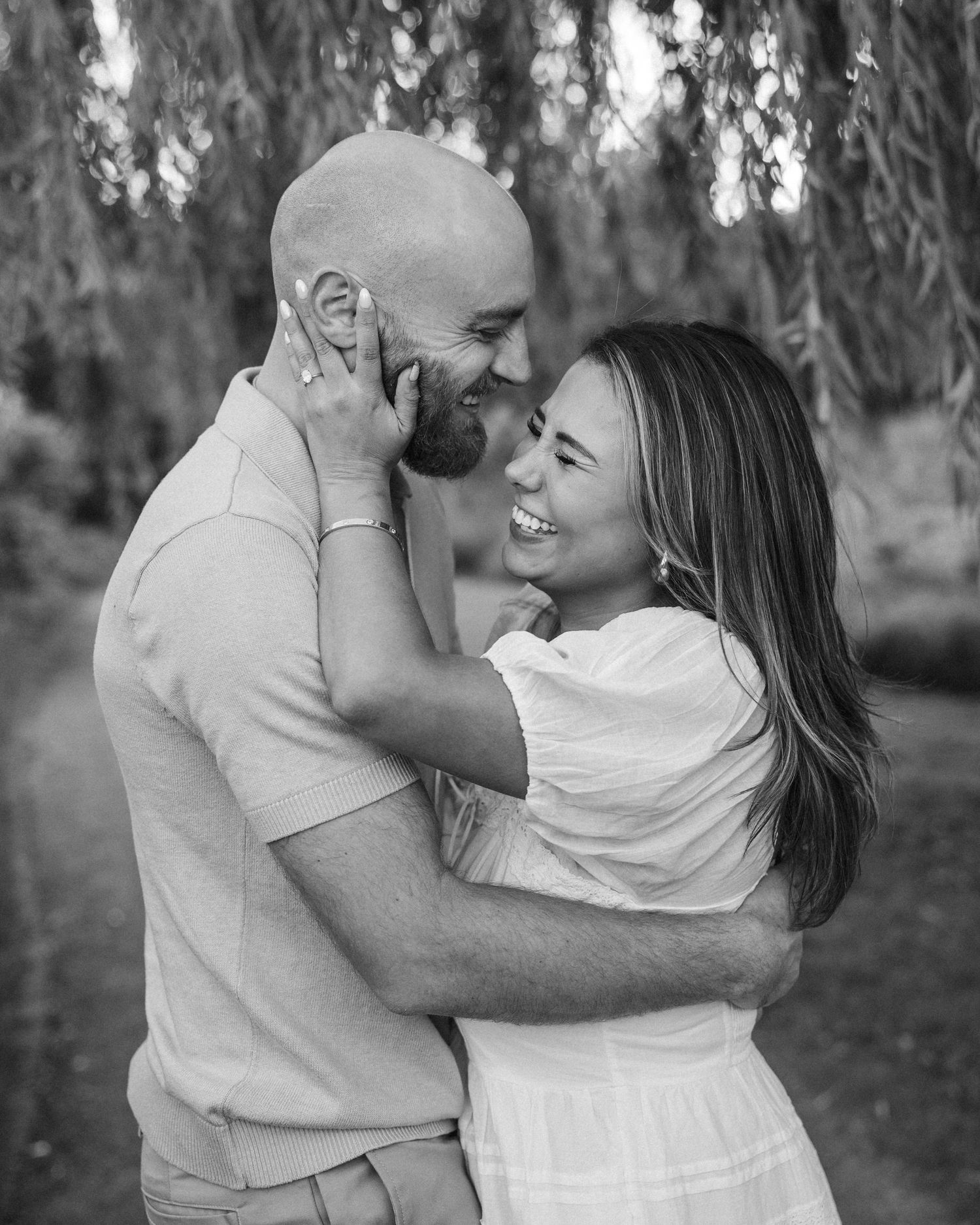 Black and white photo of a smiling couple embracing outdoors under willow tree branches.