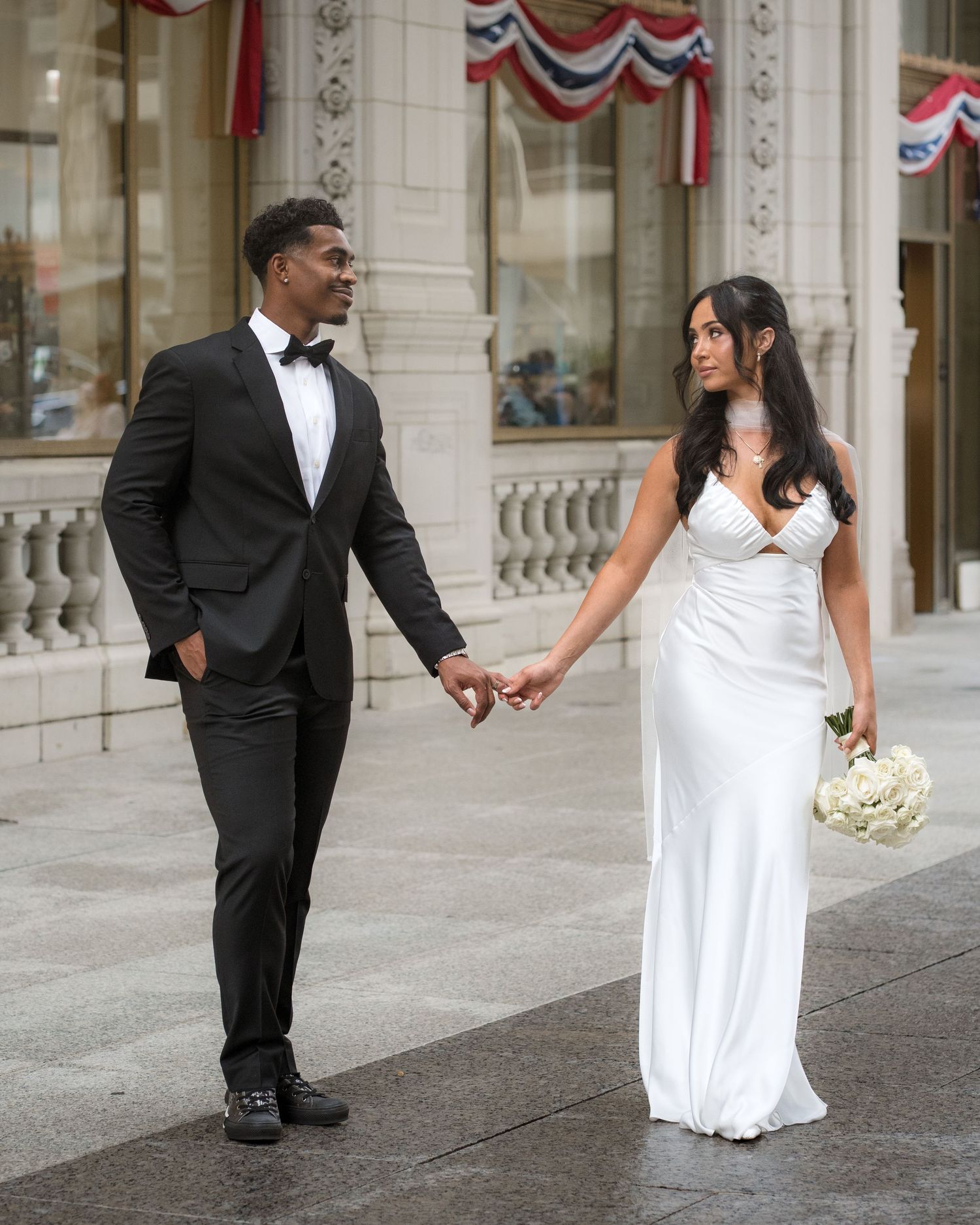 Newlyweds walking hand in hand on a city street, bride carrying white bouquet in elegant white wedding gown.