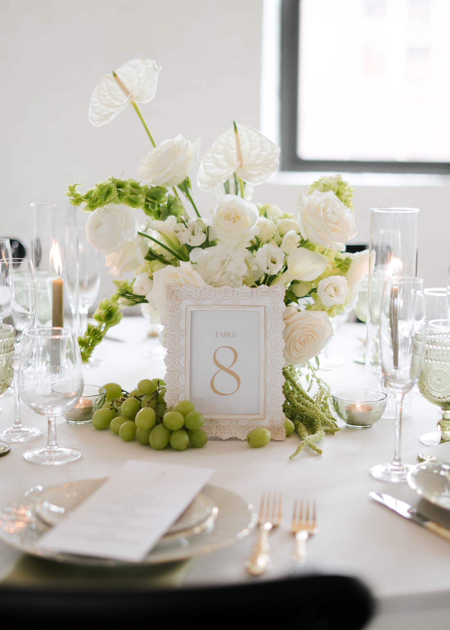 Elegant wedding table centerpiece with white ranunculus, anthurium, green grapes, and a gold table number 8 card.