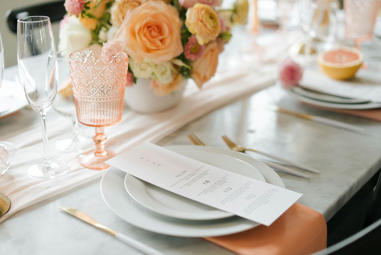 Elegant wedding table setting with peach roses, pink glassware, gold cutlery, marble surface, and a menu card on gray plates.