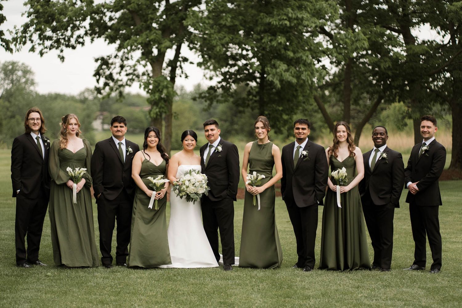 Wedding party in sage green dresses and black suits posing outdoors on green lawn with trees in background.